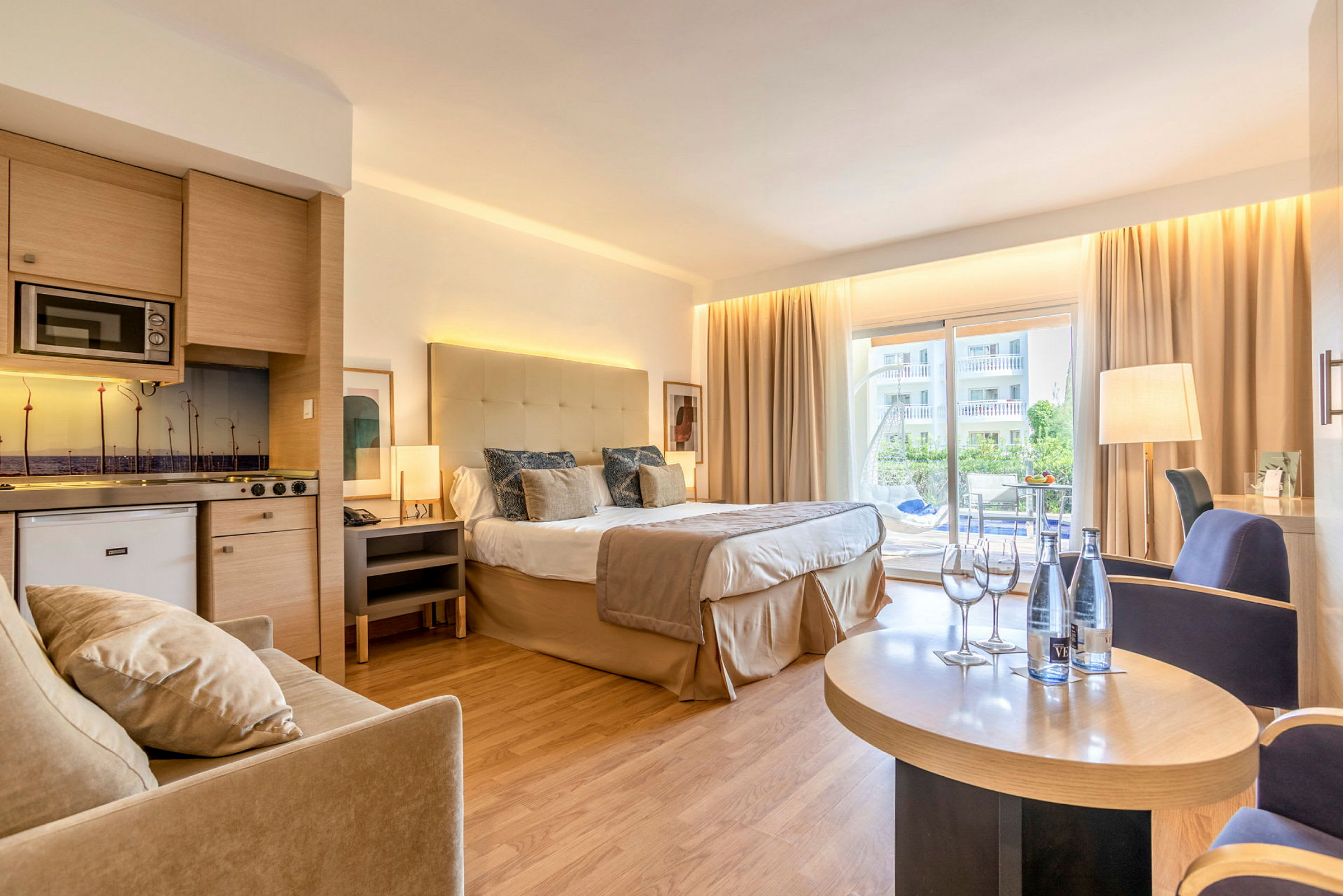 Hotellrum med dubbelsäng, sittgrupp och kök, samt balkong med utsikt. (AI-genererad text)
