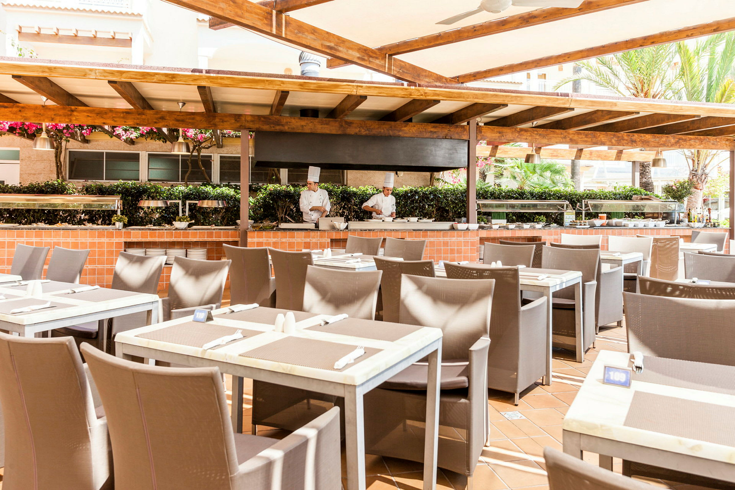 Utomhusrestaurang med dukade bord och matlagningsstation under en träpergola. (AI-genererad text)