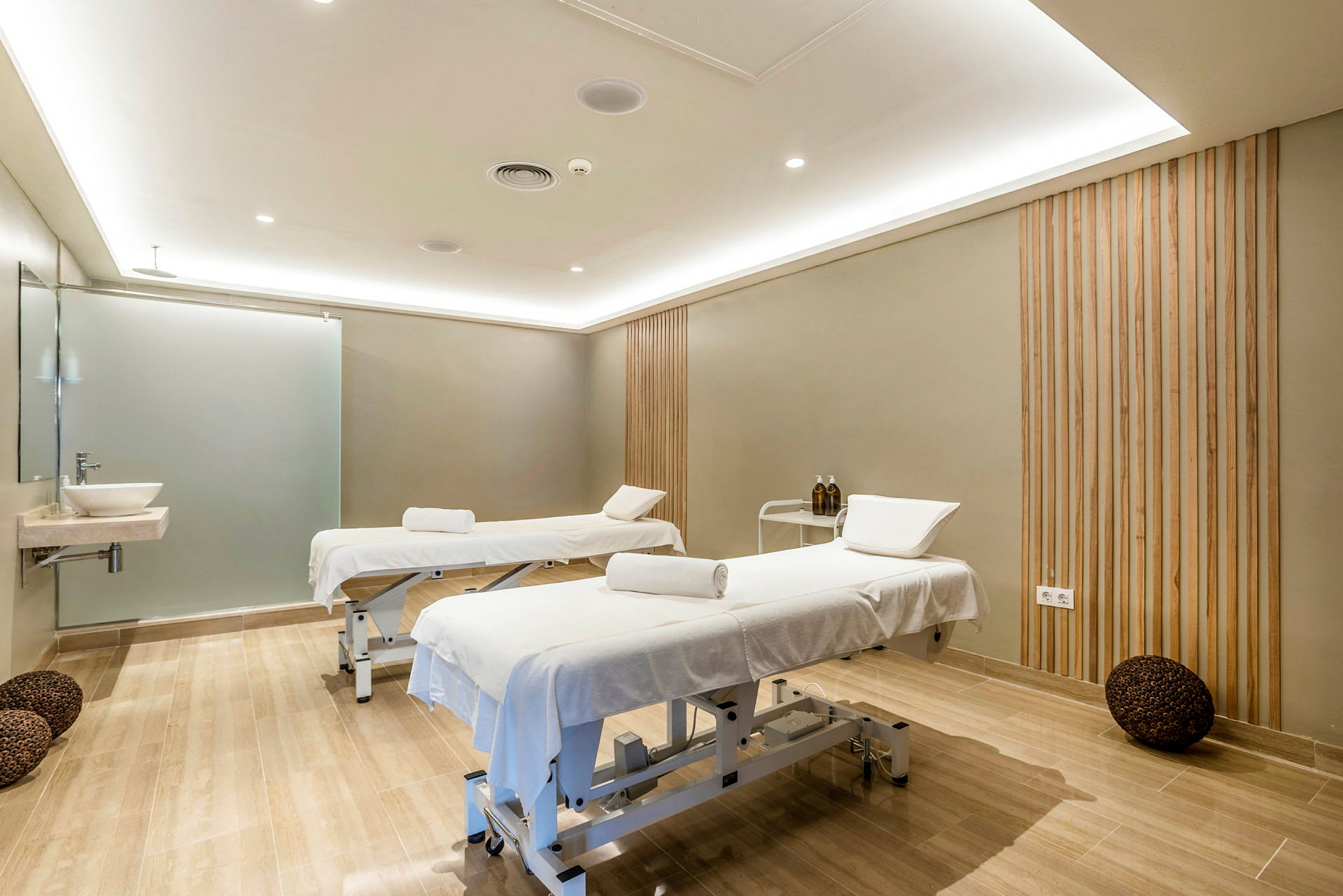 Två massagebänkar med vita handdukar och kuddar inuti ett behandlingsrum med trädetaljer på väggarna. (AI-genererad text)