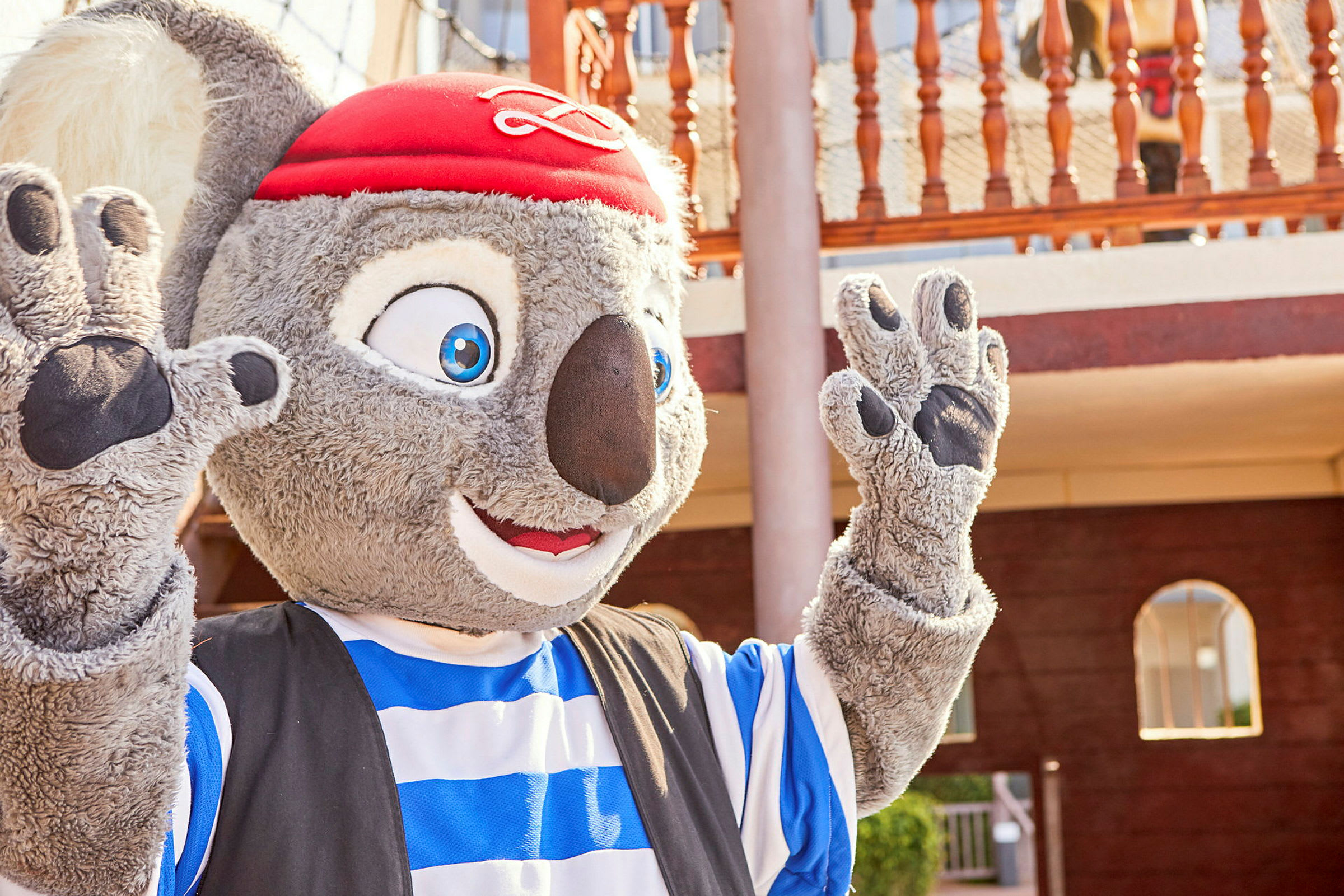 Maskot med grå päls, randig blå och vit kostym samt röd hatt, synlig framför en träbalkong. (AI-genererad text)