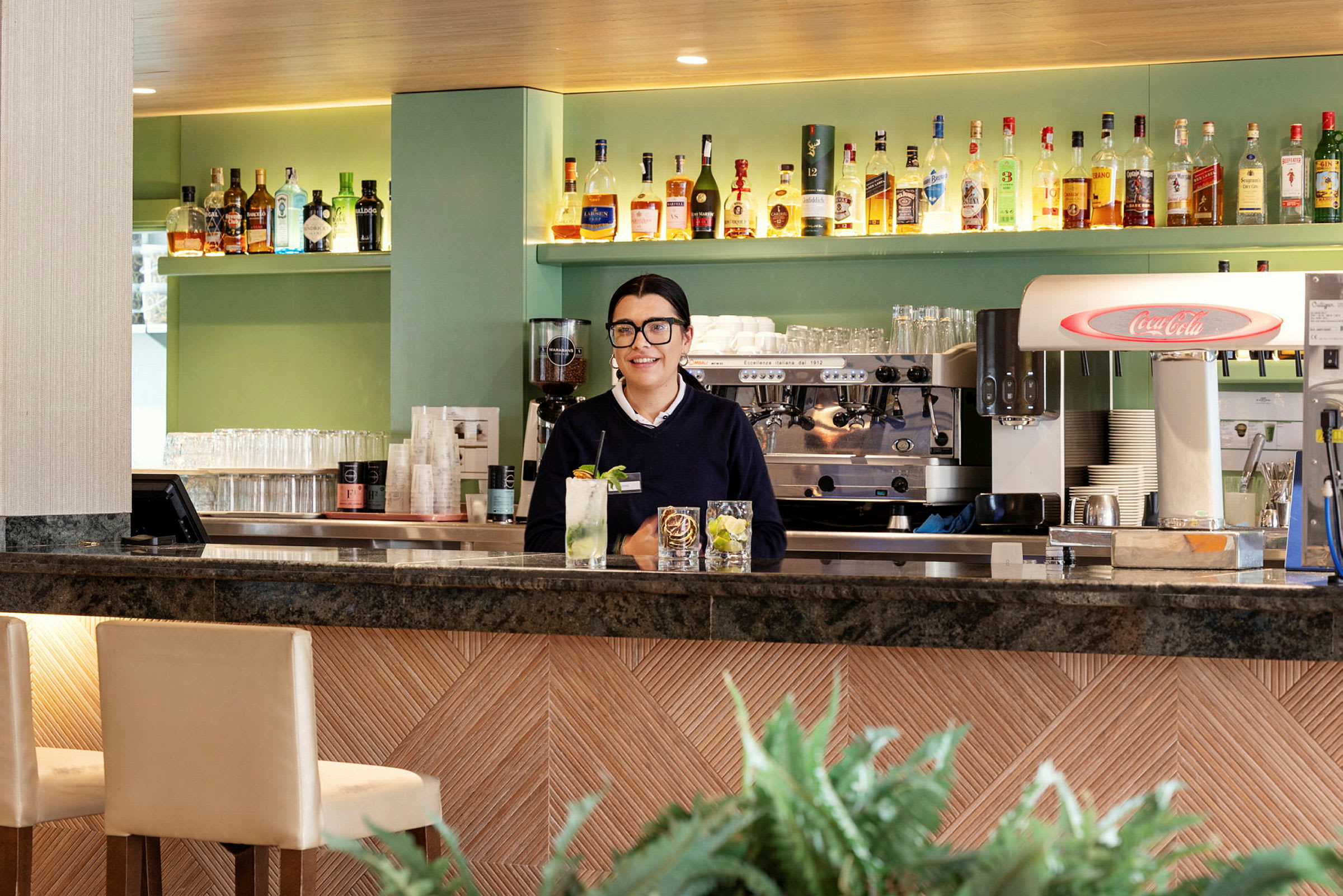 En bartender står bakom en bar med flaskor och en kaffemaskin i bakgrunden. (AI-genererad text)