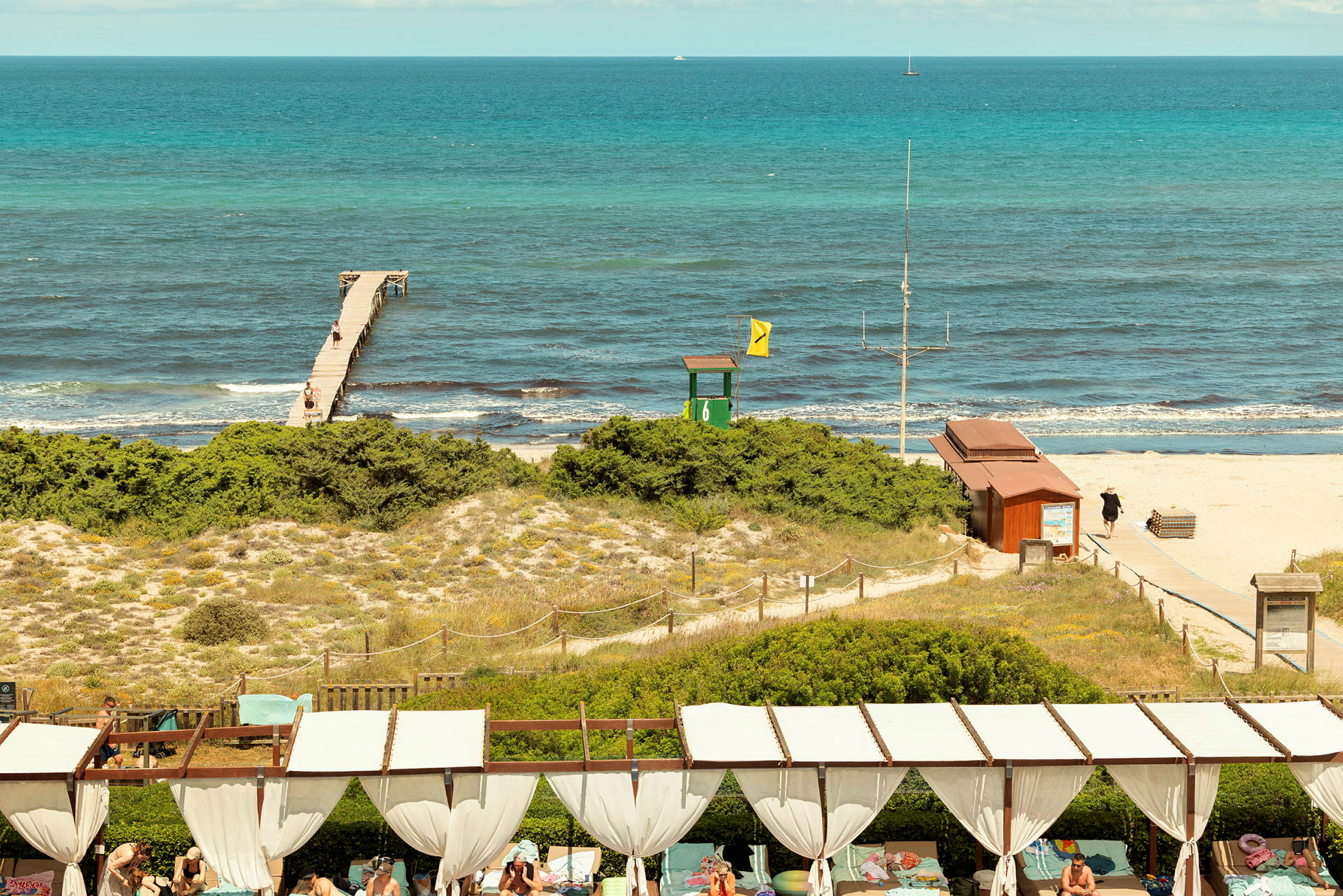 En strand med en brygga som sträcker sig ut i havet, sanddyner med vegetation i förgrunden och personer vid en relax- eller solsektion. (AI-genererad text)