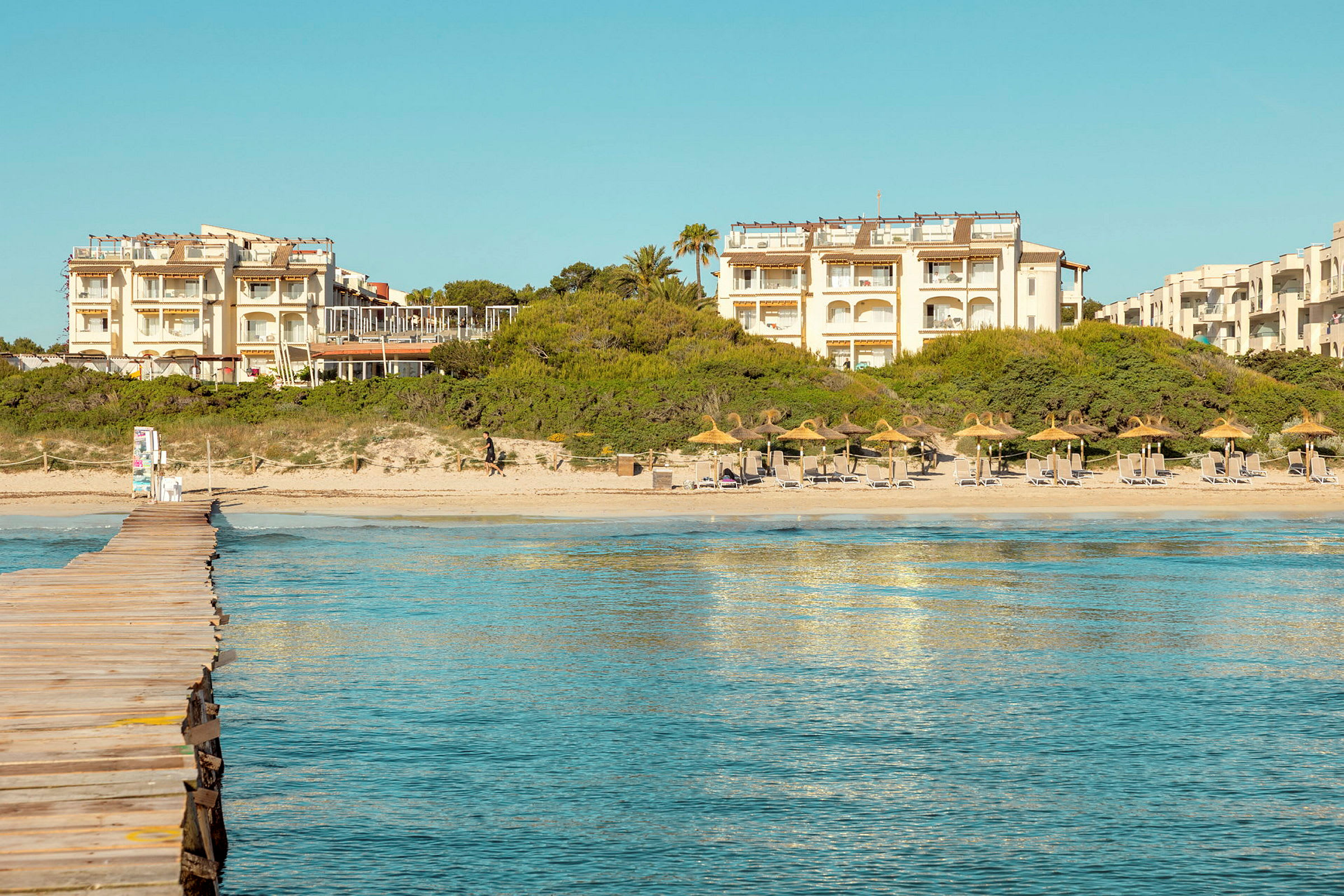 En strandpromenad leder fram till en sandstrand med parasoller och solstolar. I bakgrunden finns hotellbyggnader och grönska. (AI-genererad text)