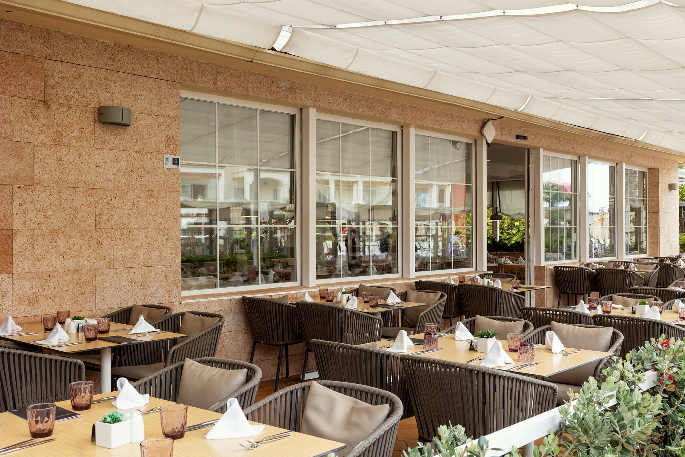 En utomhusterrass vid en restaurang med dukade bord och stolar, omgiven av gröna växter. (AI-genererad text)