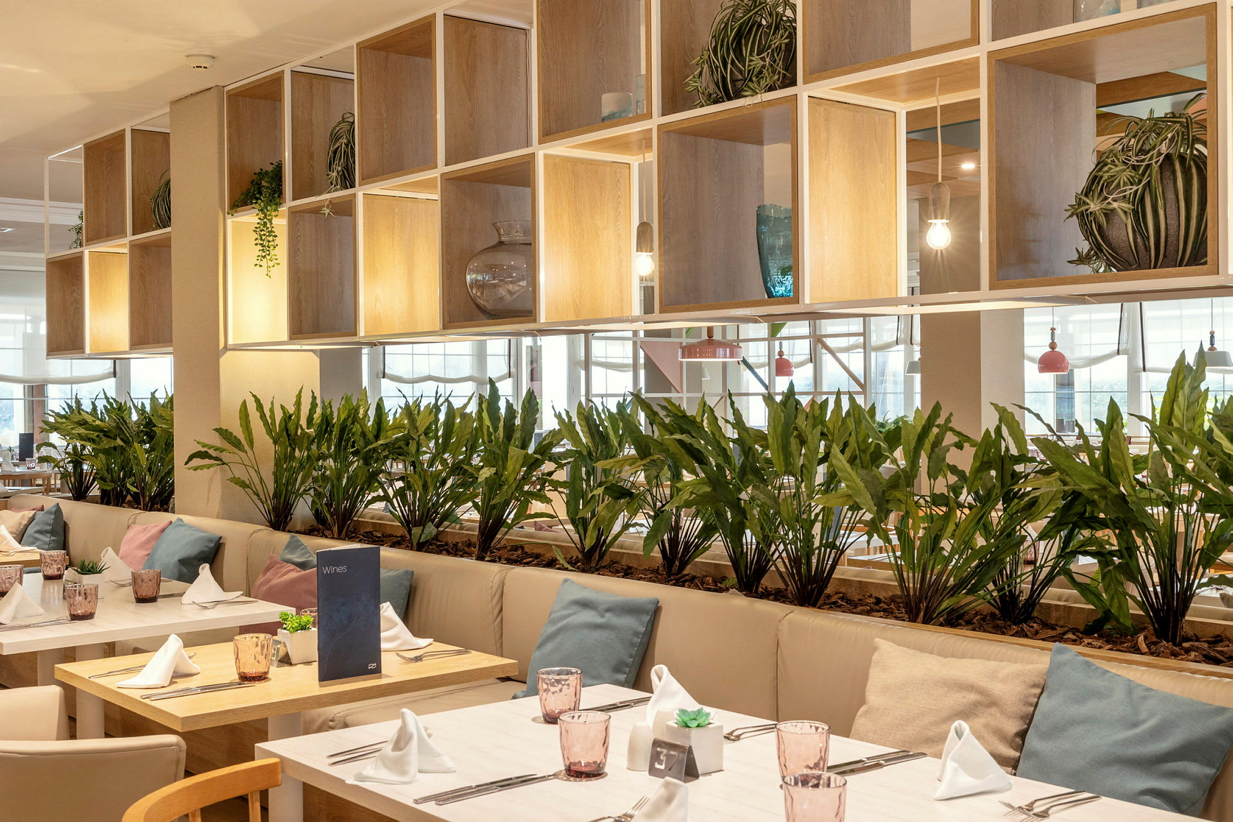 Ett modernt inrett restaurangområde med bord dekorerade med vita servetter, glas och kuddar på sofforna. Gröna växter placerade som skiljeväggar. (AI-genererad text)