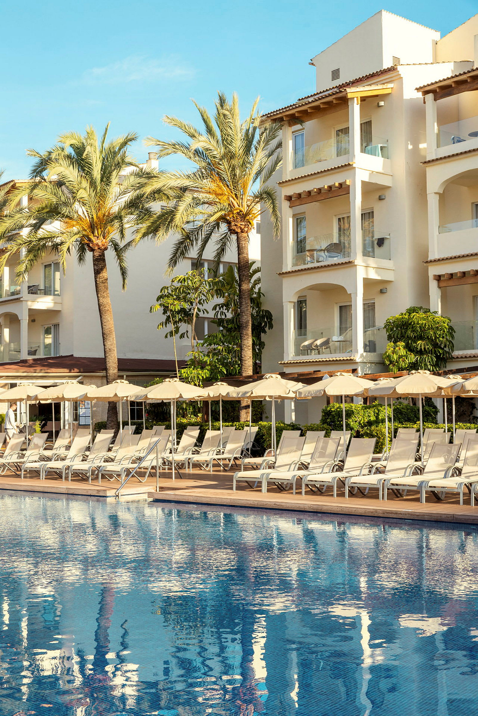 Bild föreställer en utomhuspool med solstolar och parasoll framför en hotellbyggnad med balkonger. (AI-genererad text)