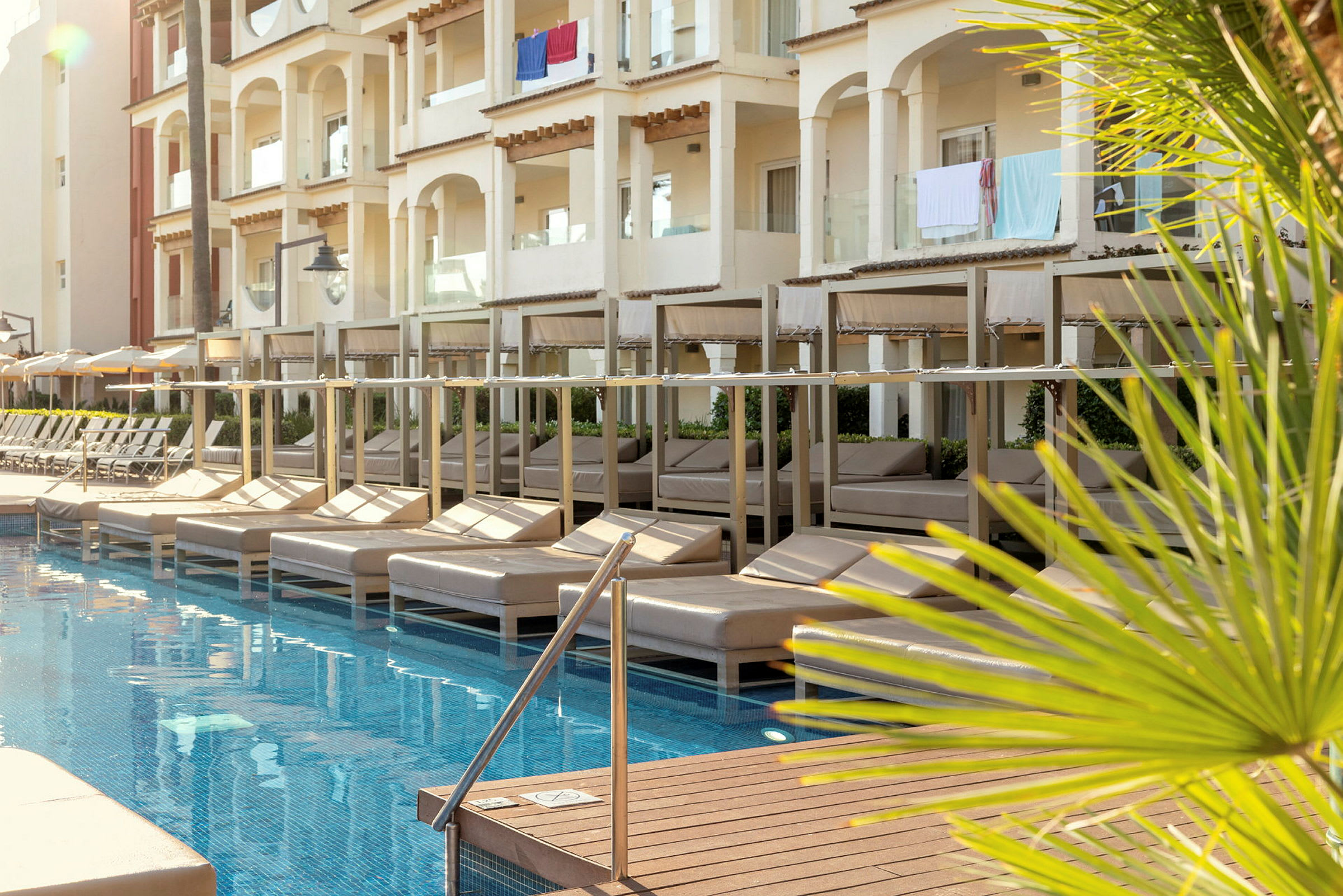 En pool med solbäddar under skuggtak vid ett hotell med balkonger. (AI-genererad text)