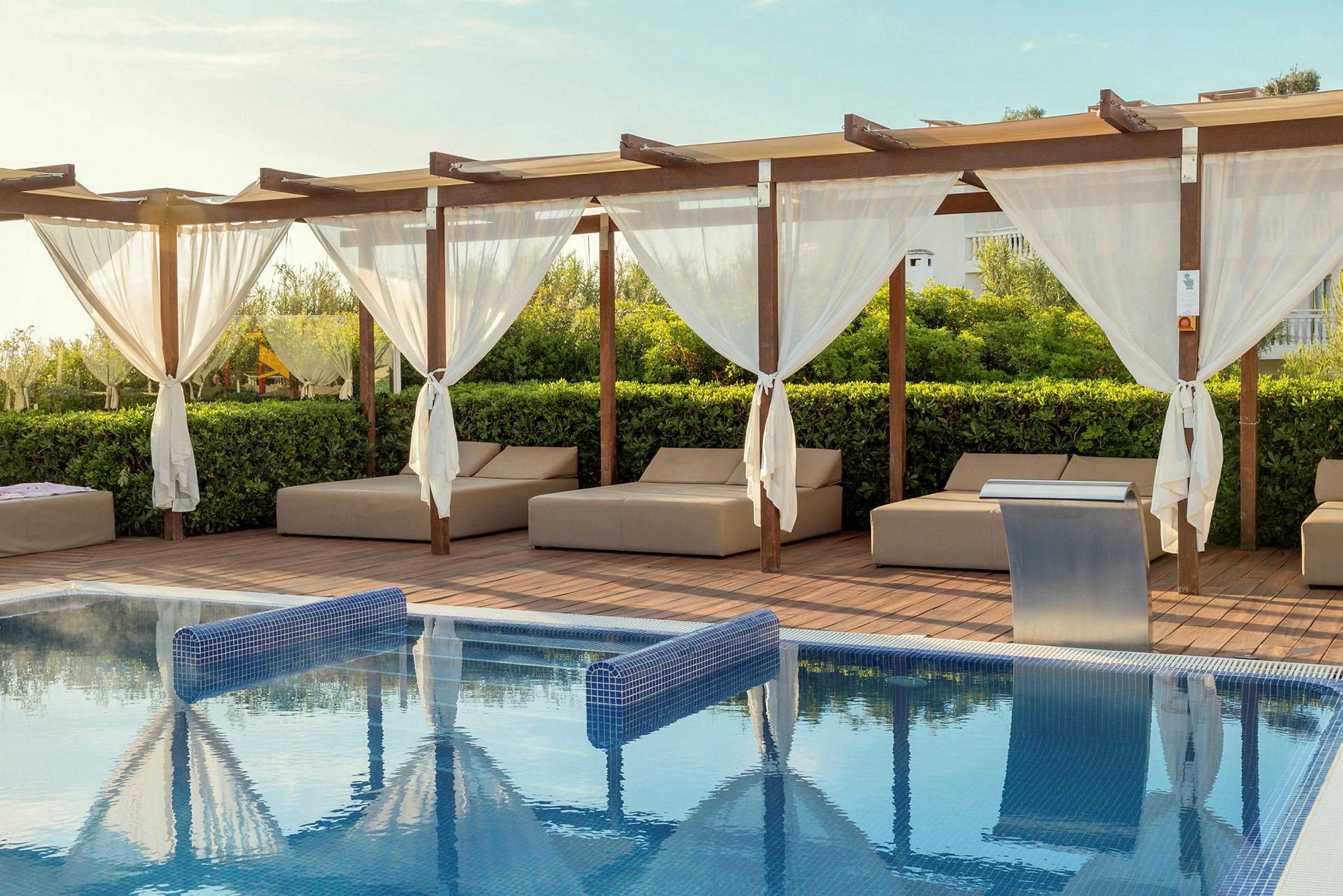 En pool med blått vatten, omgiven av solstolar och en trall med pergola och vita gardiner. (AI-genererad text)