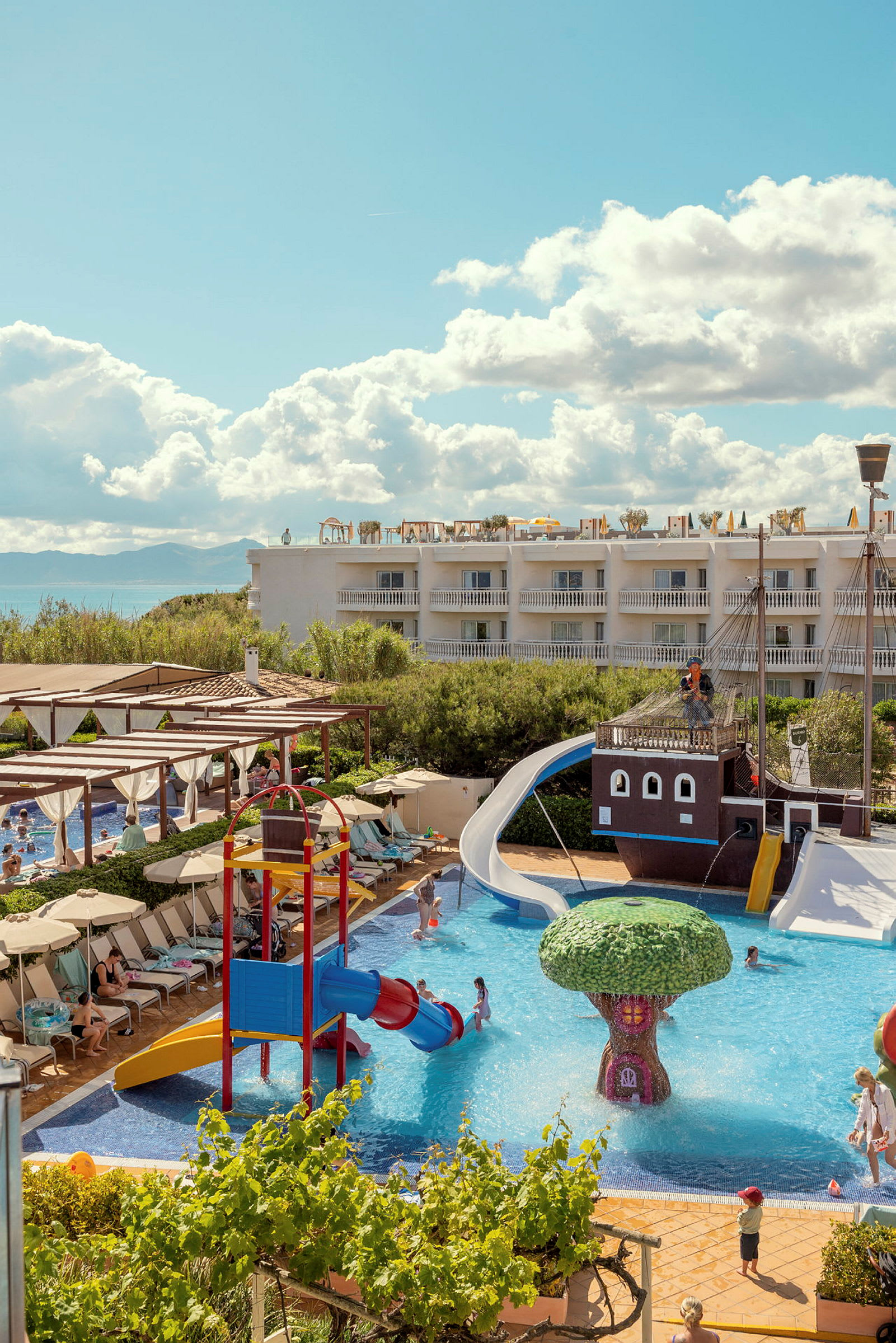 Bild av en resort med poolområde, barn leker vid vattenrutschbanor och simmar nära en fontän formad som ett träd. (AI-genererad text)