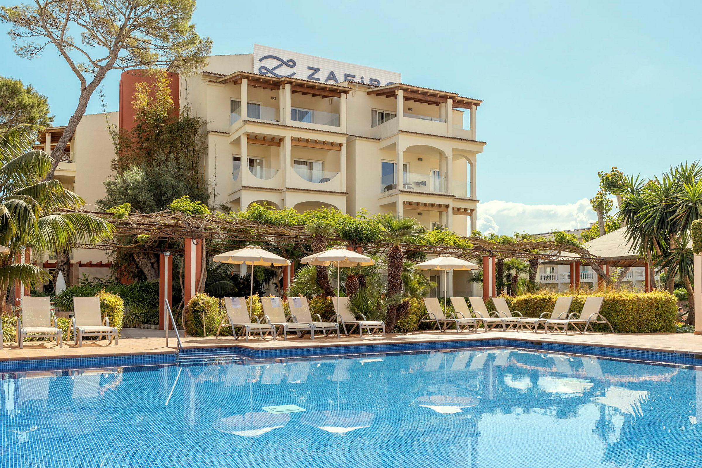 Ett hotell med en pool i förgrunden, omringat av solstolar och parasoller. Byggnaden omges av grönska och har träbjälkar och pelare på balkongerna. (AI-genererad text)