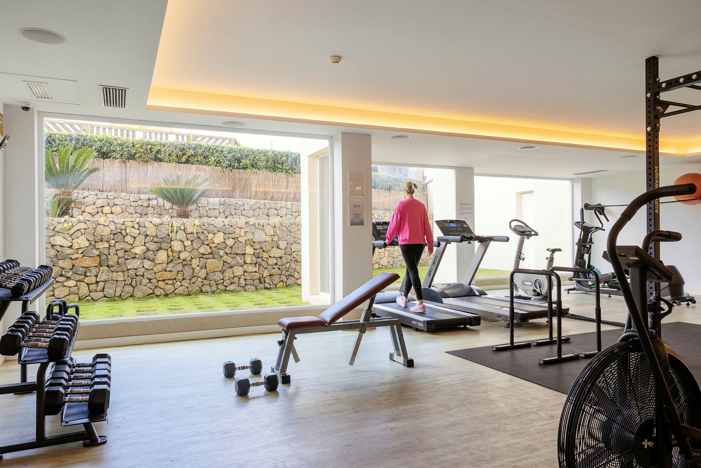 Ett gym med träningsutrustning såsom löpband, vikter och träningscykel. Någon går på ett löpband. (AI-genererad text)