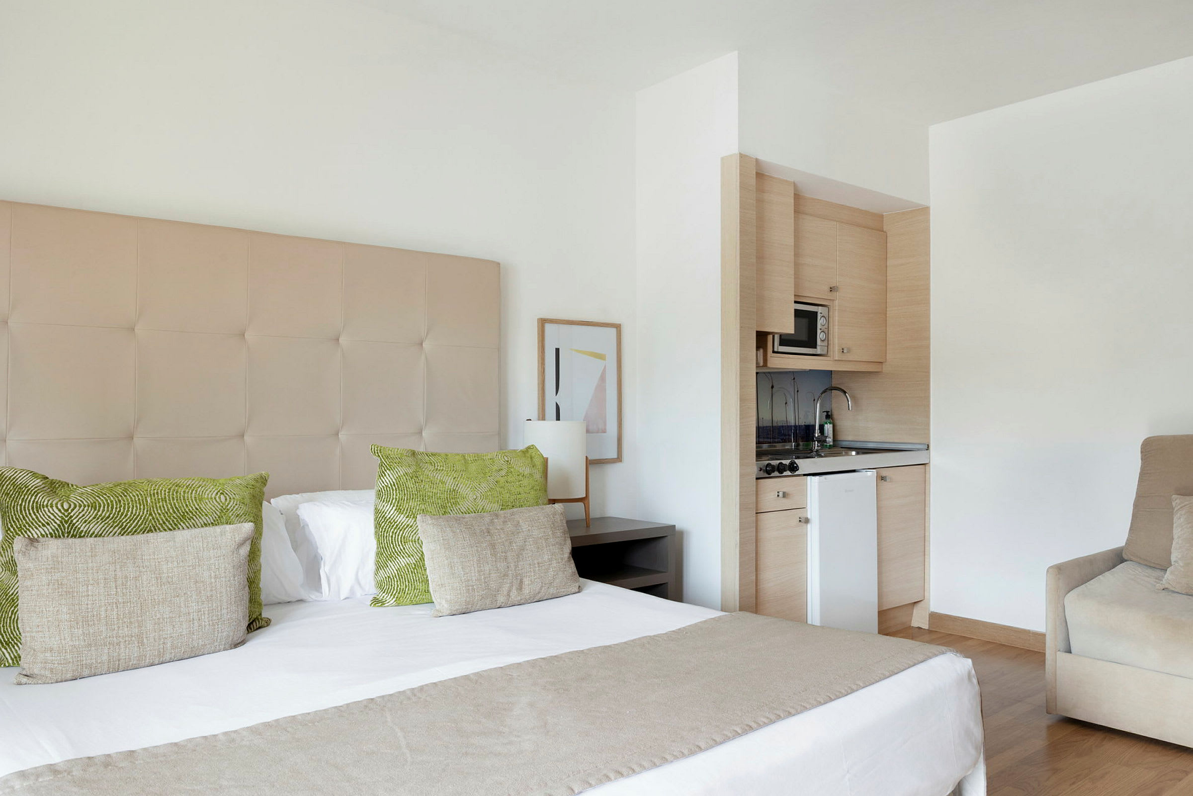 Ett hotellrum med en dubbelsäng, gröna och beige kuddar, en liten köksdel med mikrovågsugn och diskho samt ett beige fåtölj. (AI-genererad text)