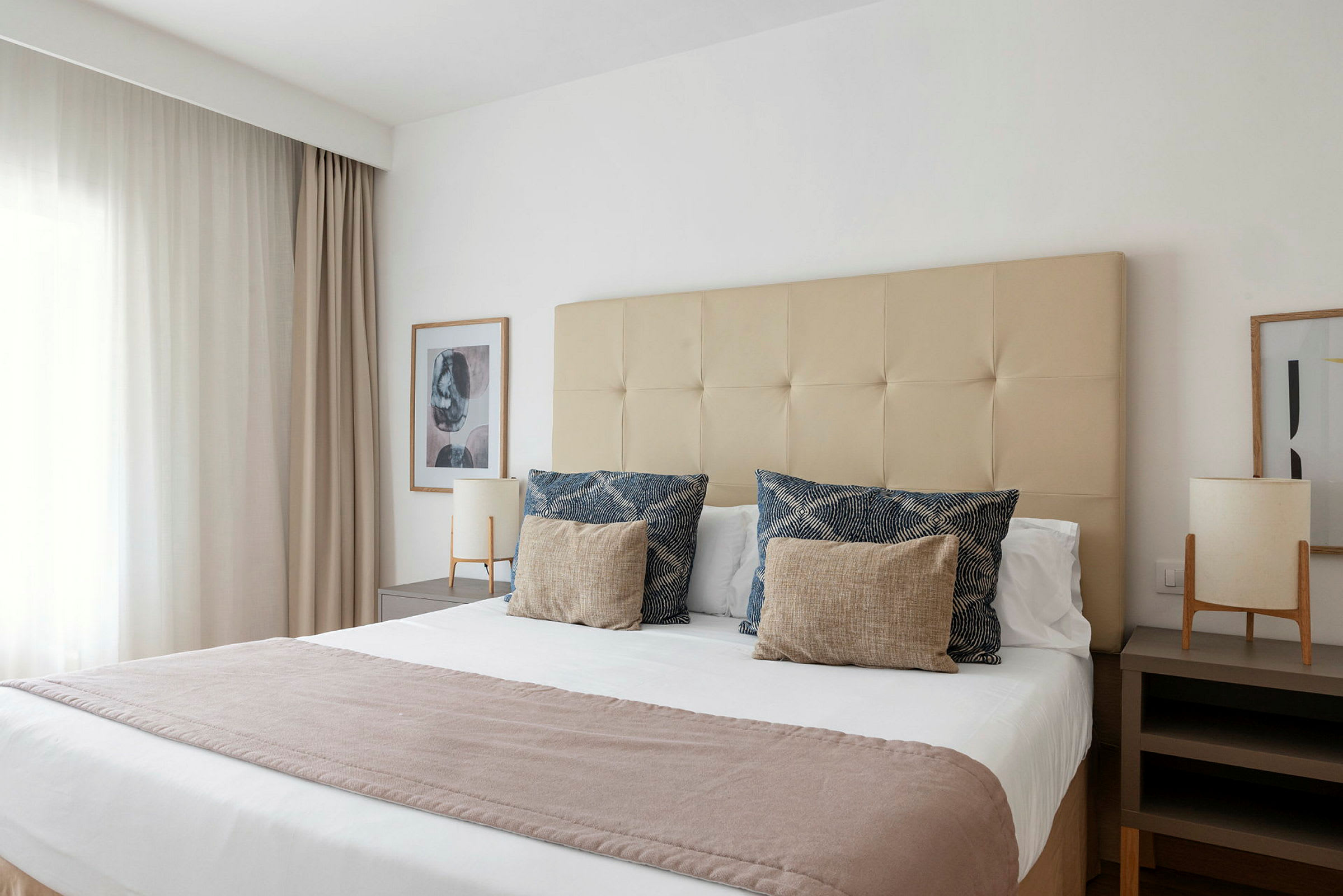 En hotellrum med en stor säng täckt av vita lakan, dekorativa kuddar och en ljus filt. Sängen är placerad mellan två sängbord med lampor. (AI-genererad text)