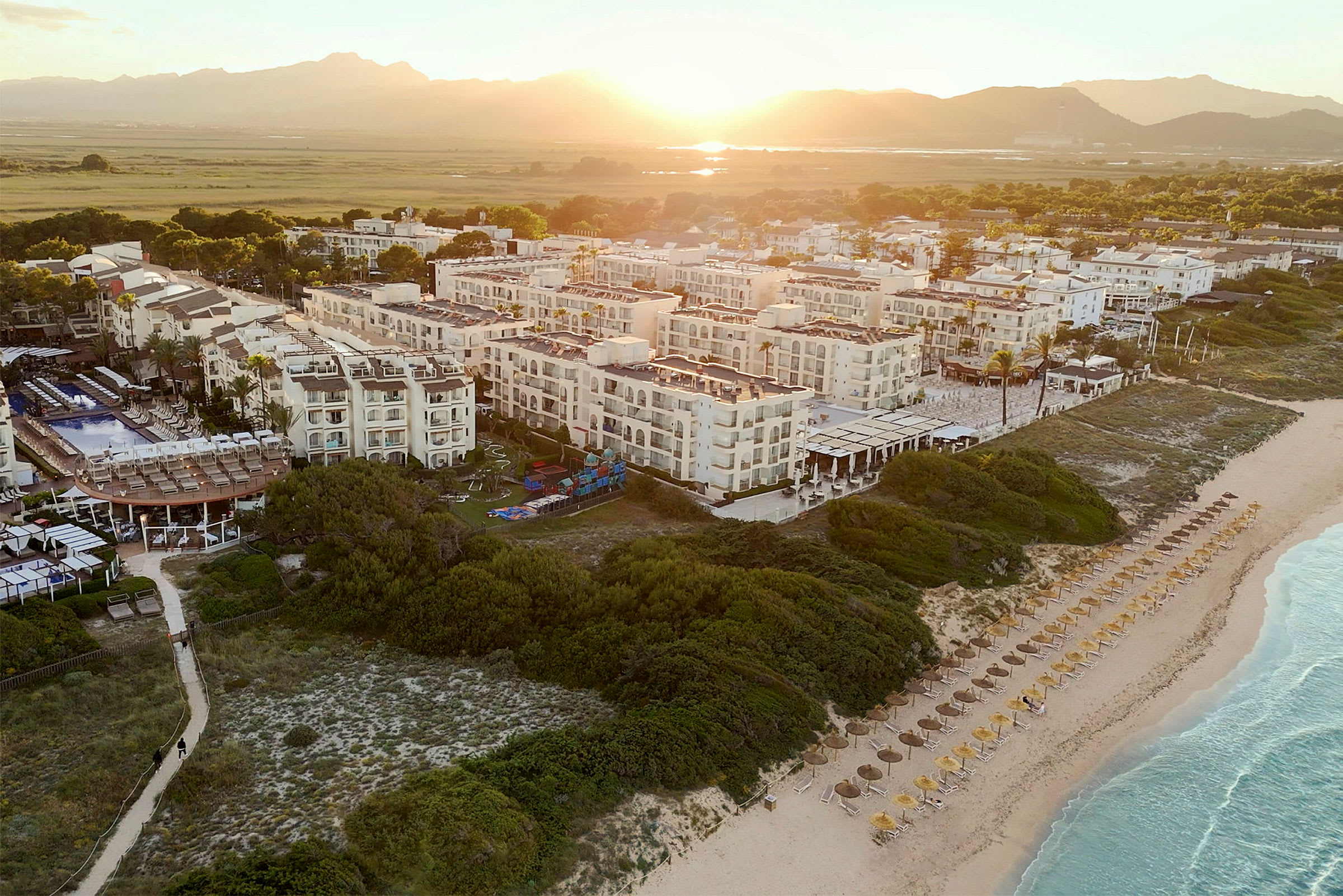 Flygbild av ett hotellkomplex vid stranden, omgiven av grönska och med soluppgång i bakgrunden. (AI-genererad text)