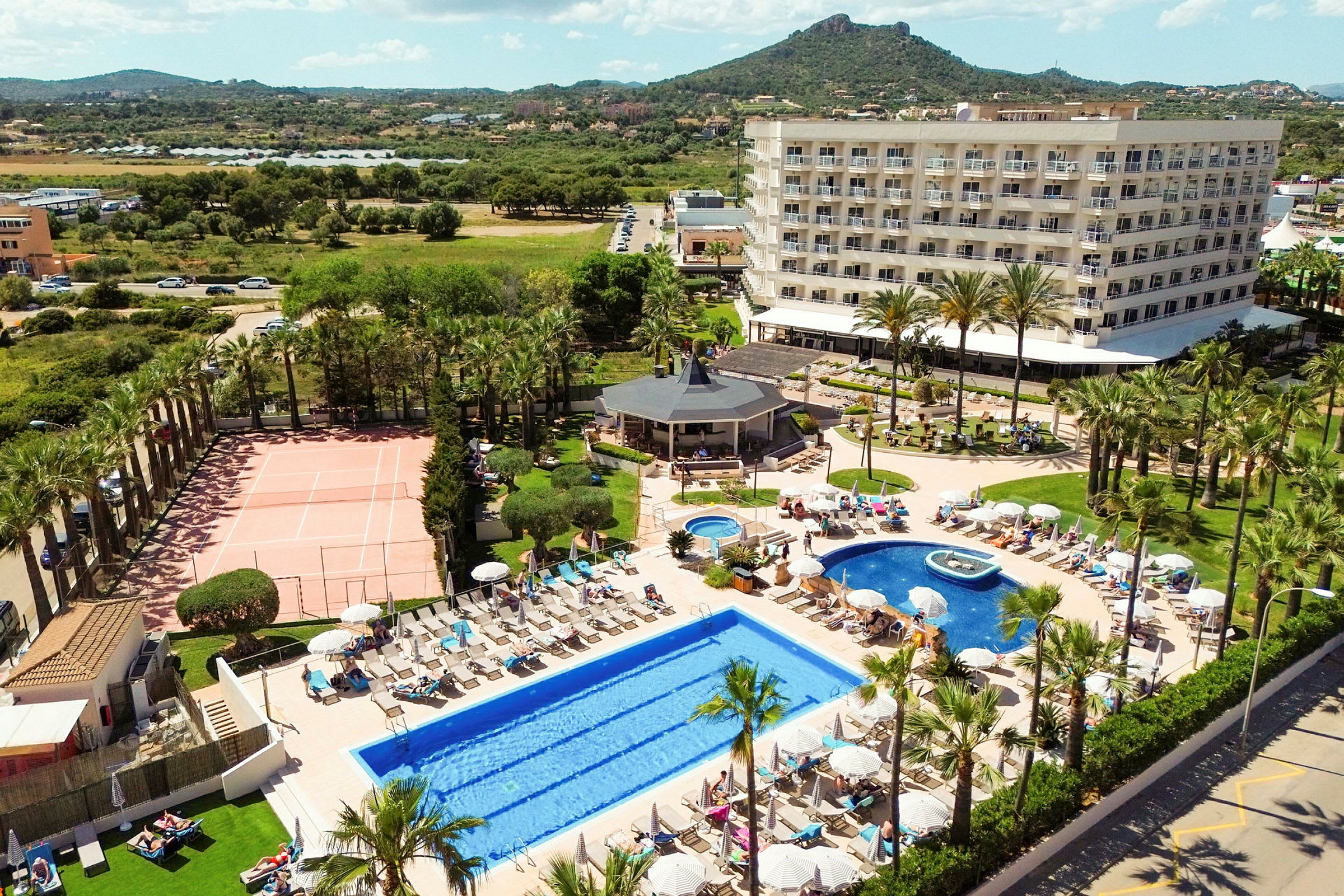 Hotellkomplex med pooler, solstolar, tennisplan och trädgård i förgrunden, samt omgivande grönska och berg i bakgrunden. (AI-genererad text)