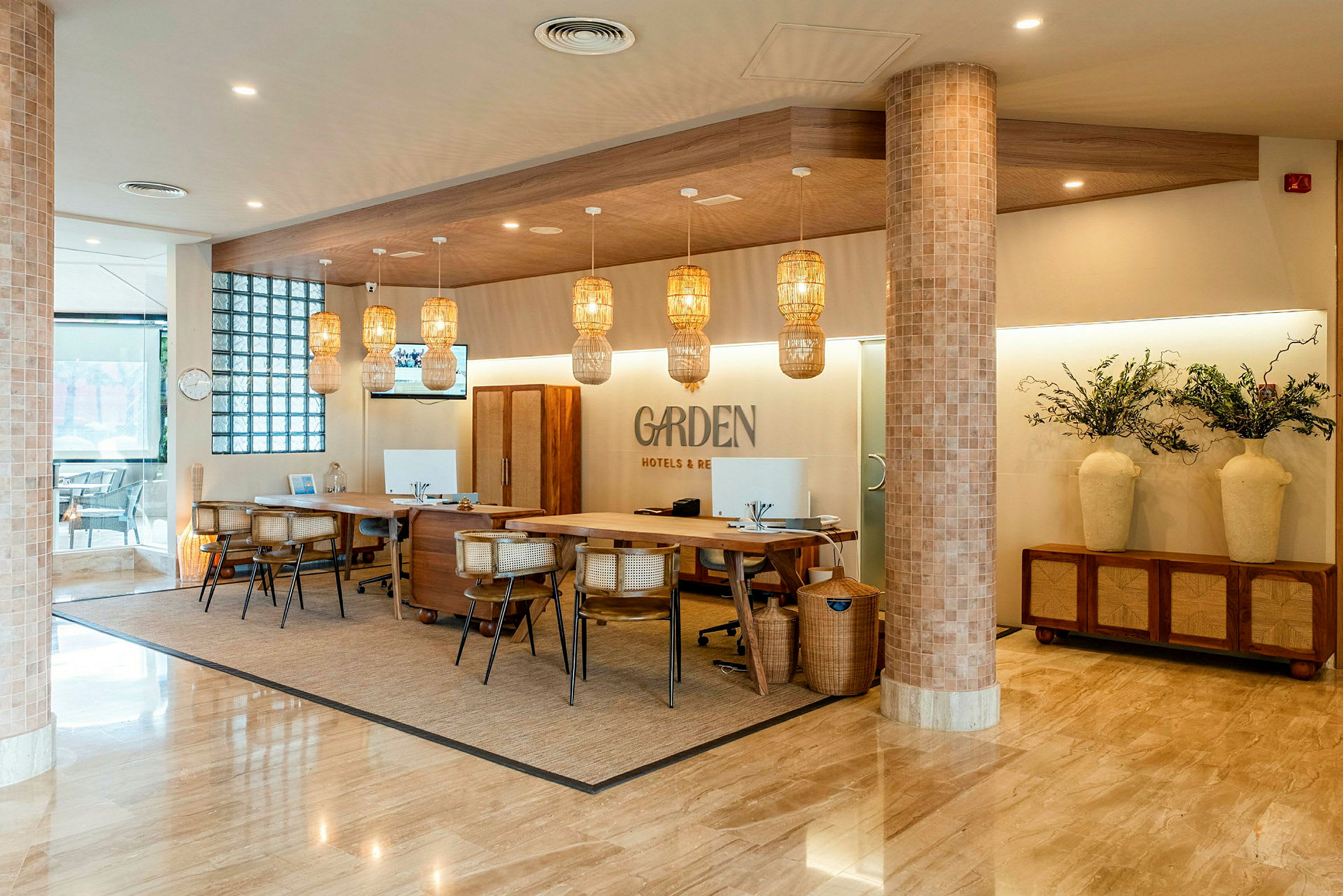 Bilden visar en hotellreception med flera skrivbord utrustade med stolar och datorer. Taklampor av rotting hänger från taket. Väggar och golv har en modern, ljus design. (AI-genererad text)