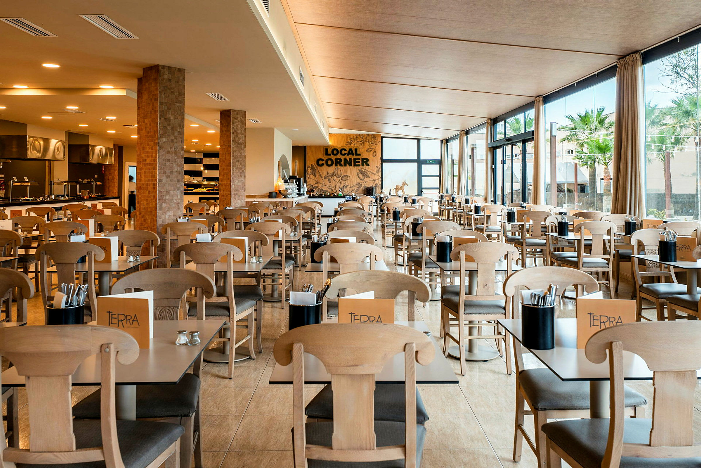 Ett ljust och rymligt restaurangområde med bord och stolar i trä. Stora fönster med utsikt mot grönska. (AI-genererad text)