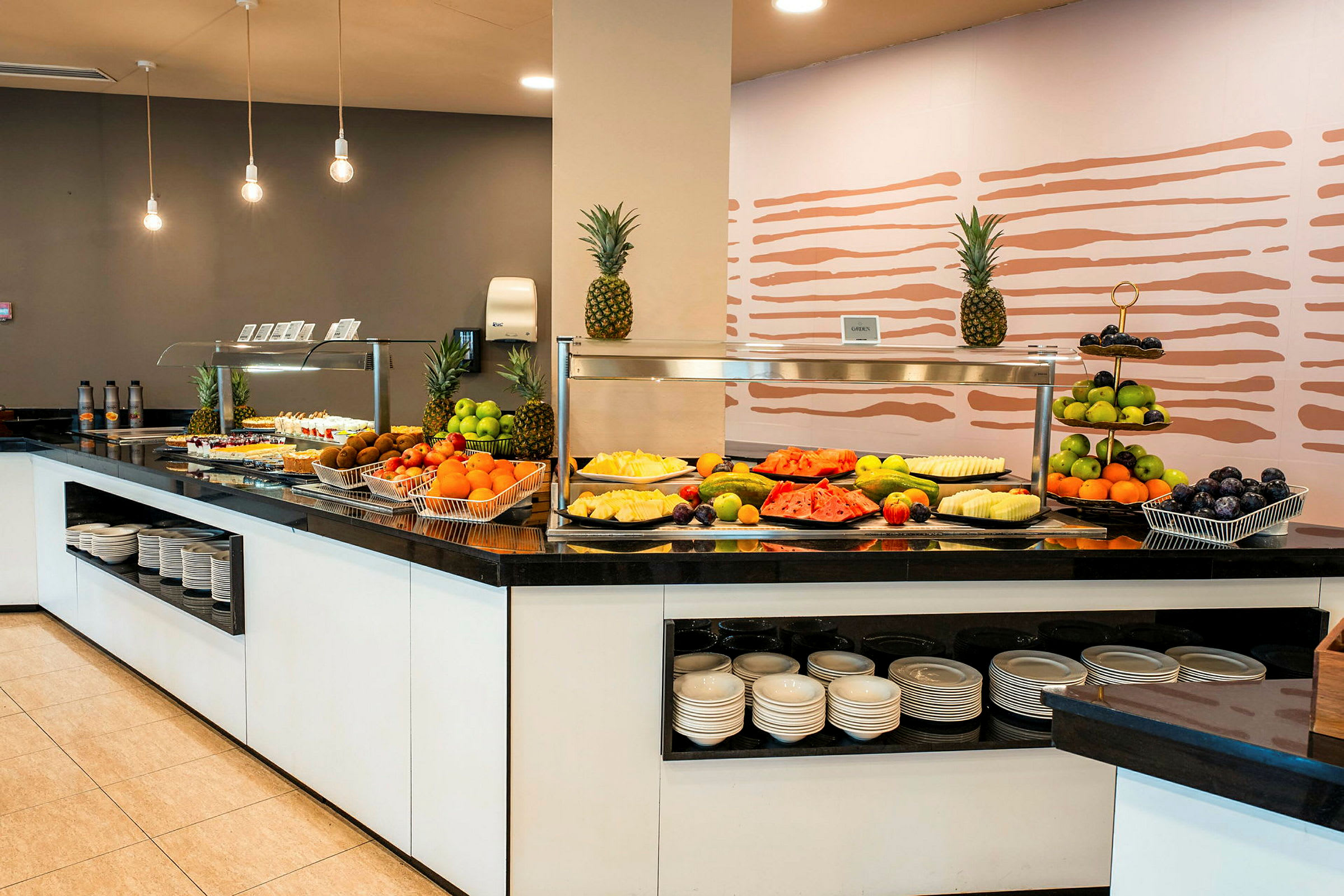 Bild på en buffédisk med färsk frukt och mat upplagd, under belysning med moderna taklampor. (AI-genererad text)