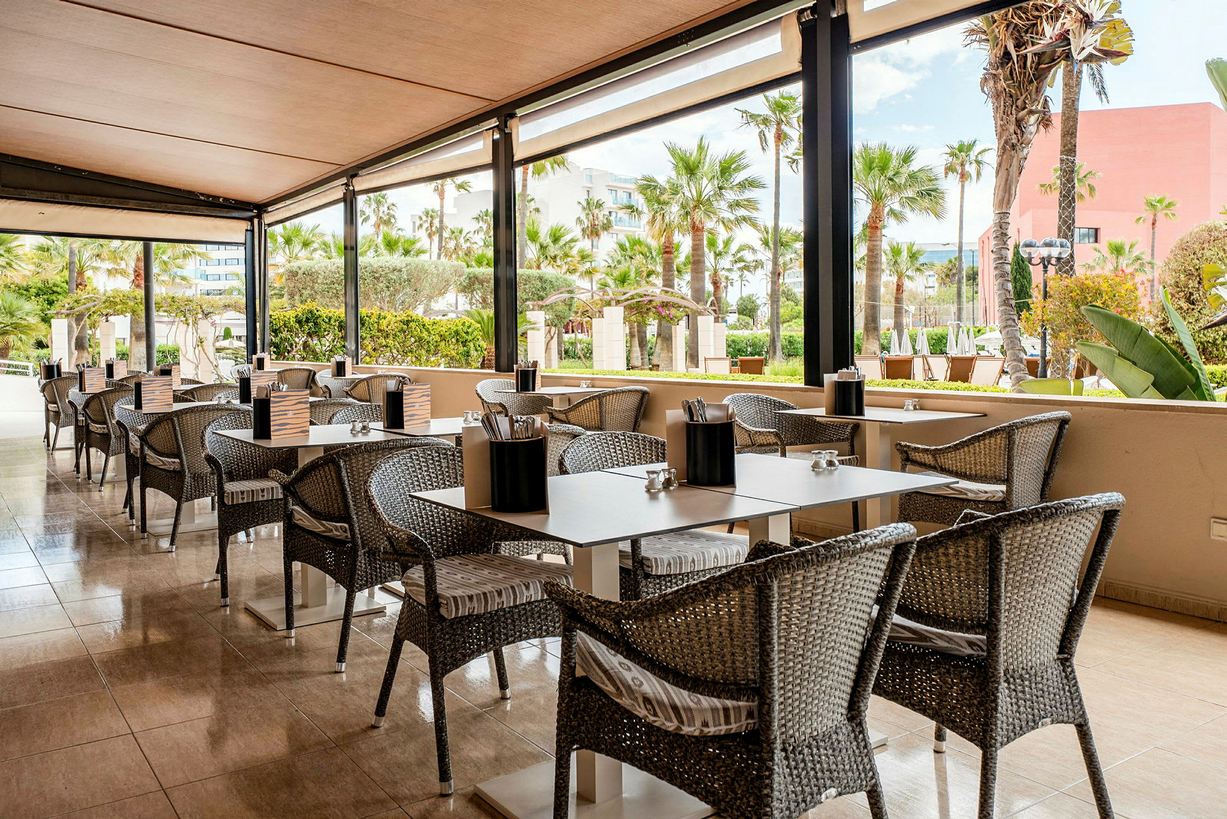En utomhusrestaurang med bord och stolar på en terrass med utsikt över en trädgård med palmer. (AI-genererad text)