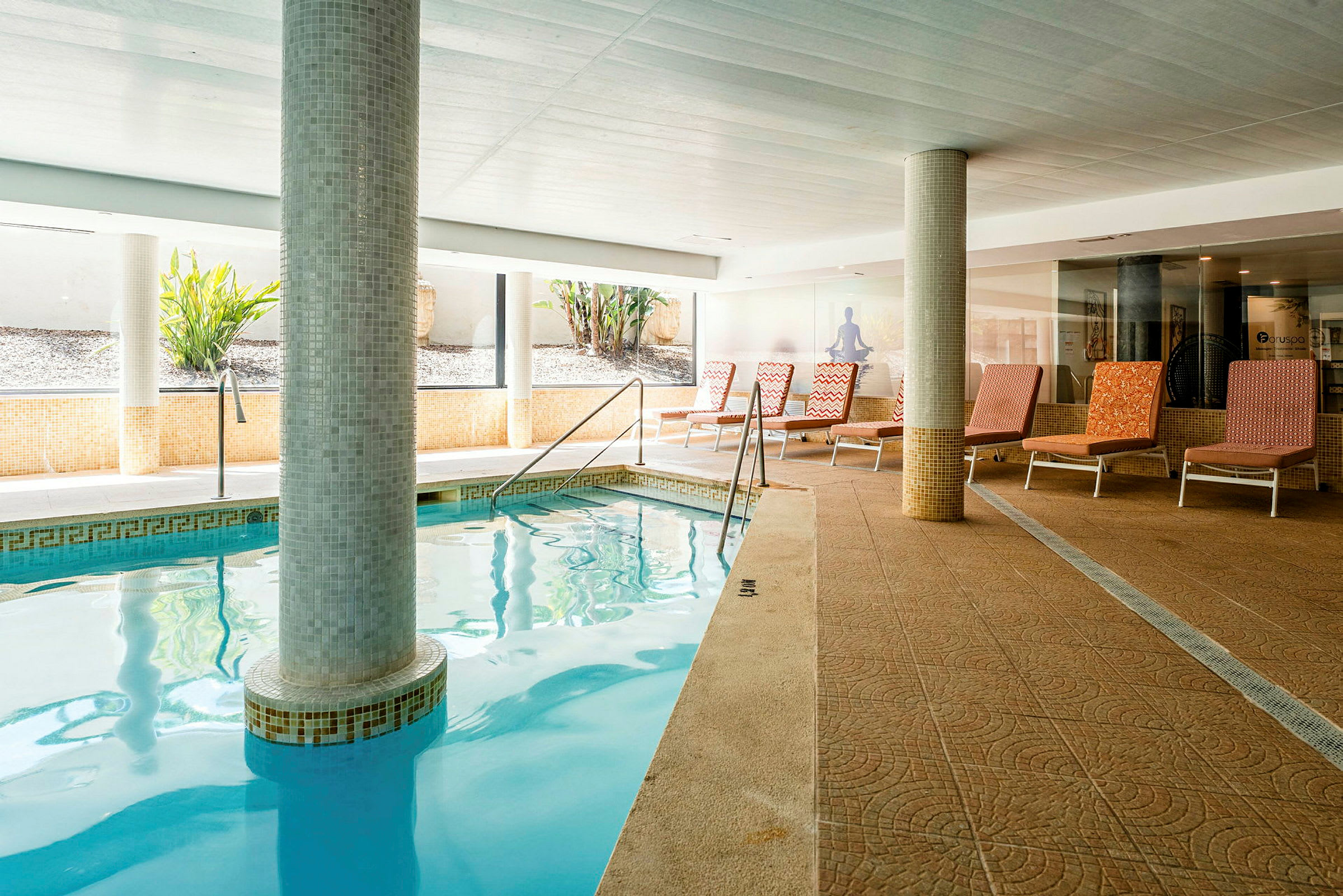 En inomhuspool med mosaikklädsel och en bred kant där solstolar står uppradade. (AI-genererad text)