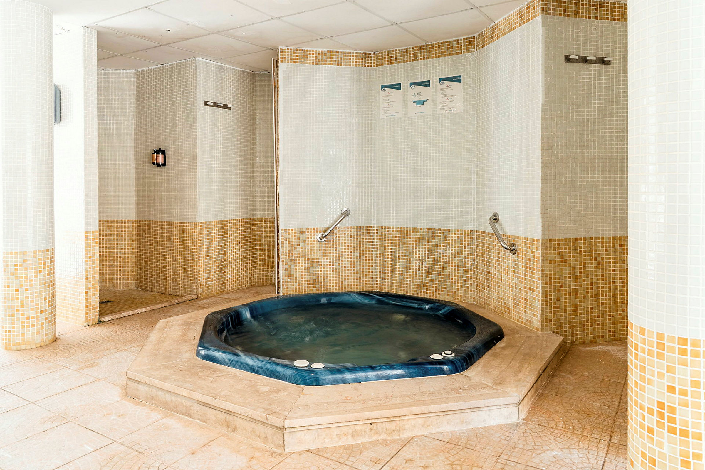 Bilden visar ett rum med en bubbelpool omgiven av mosaikdekorerade väggar. Bubbelpoolen är blå och nedsänkt i golvet. (AI-genererad text)