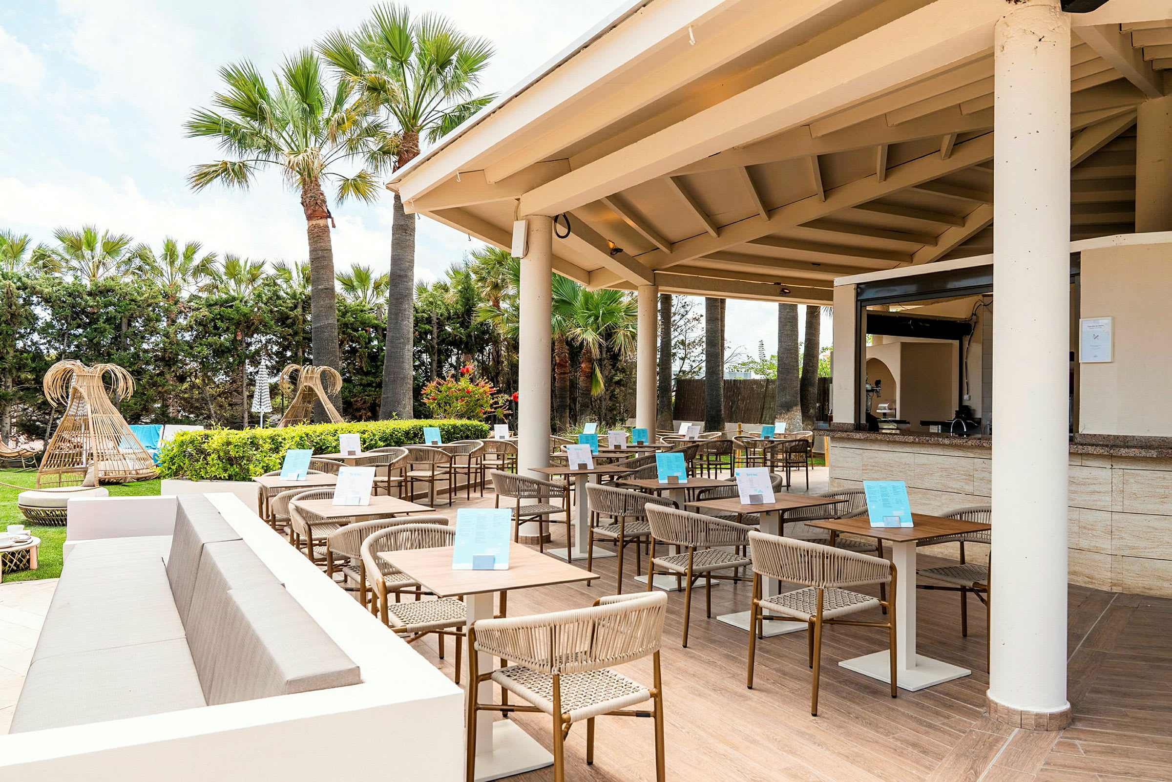 En utomhusrestaurang med bord och stolar under ett tak, omgiven av växter och palmer. (AI-genererad text)
