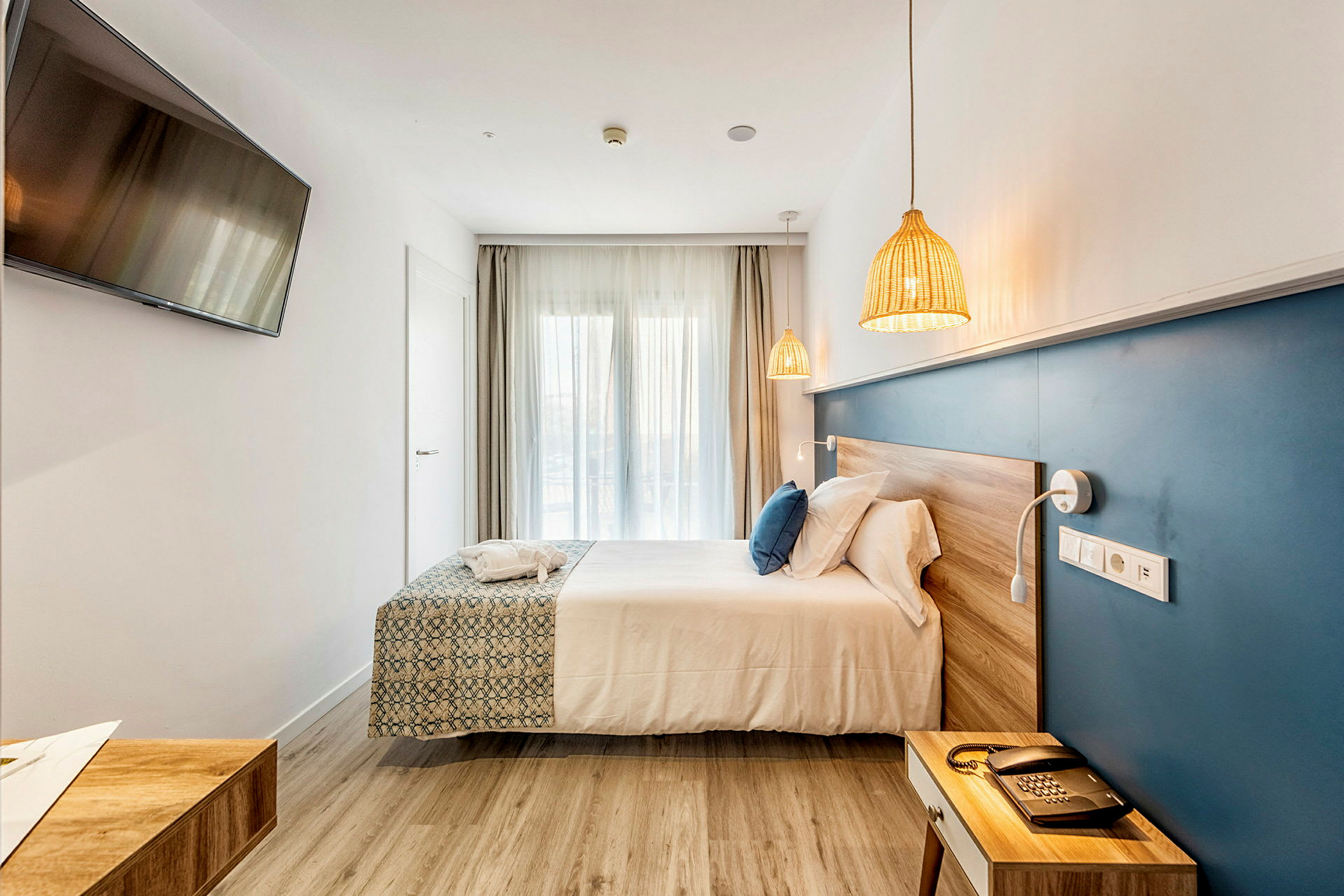Ett hotellrum med en dubbelsäng, en blå väggpanel, en TV på väggen och naturliga träytor. (AI-genererad text)