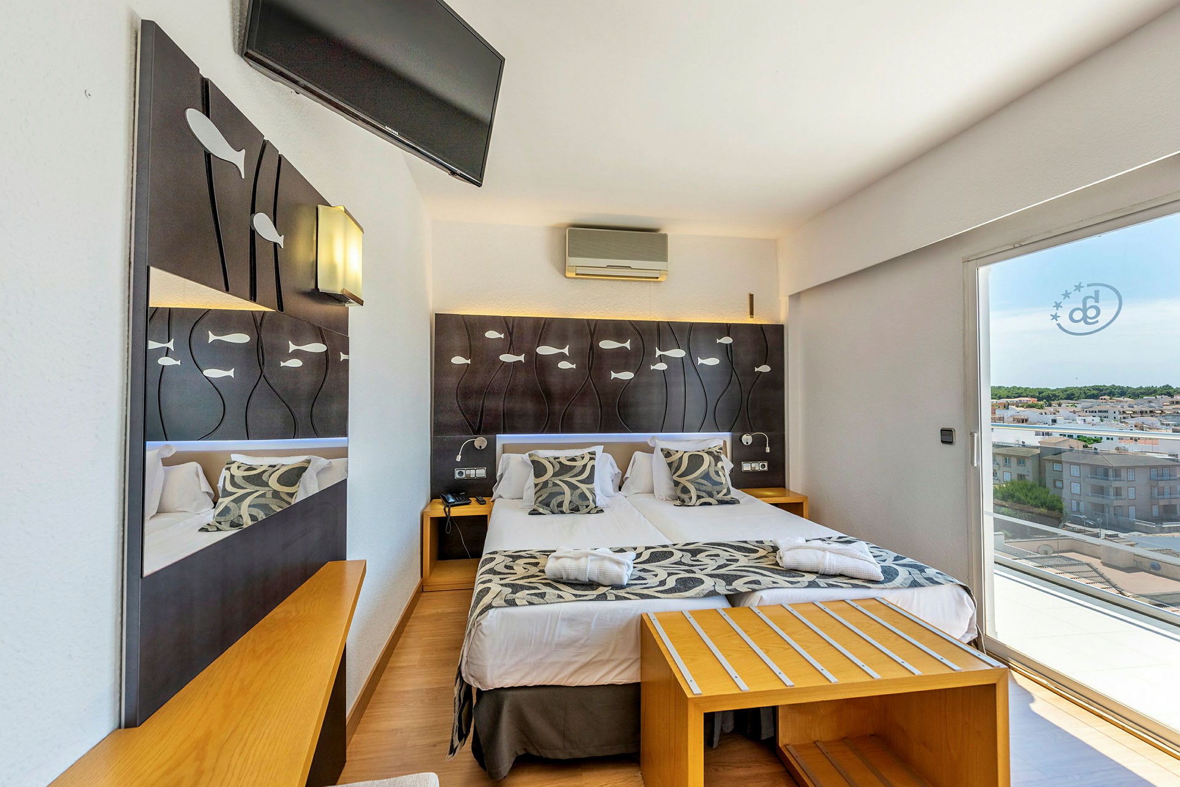 Hotellrum med dubbelsäng, dekorerat med fiskmotiv på sänggaveln och en utsikt mot staden genom glasdörrar. (AI-genererad text)