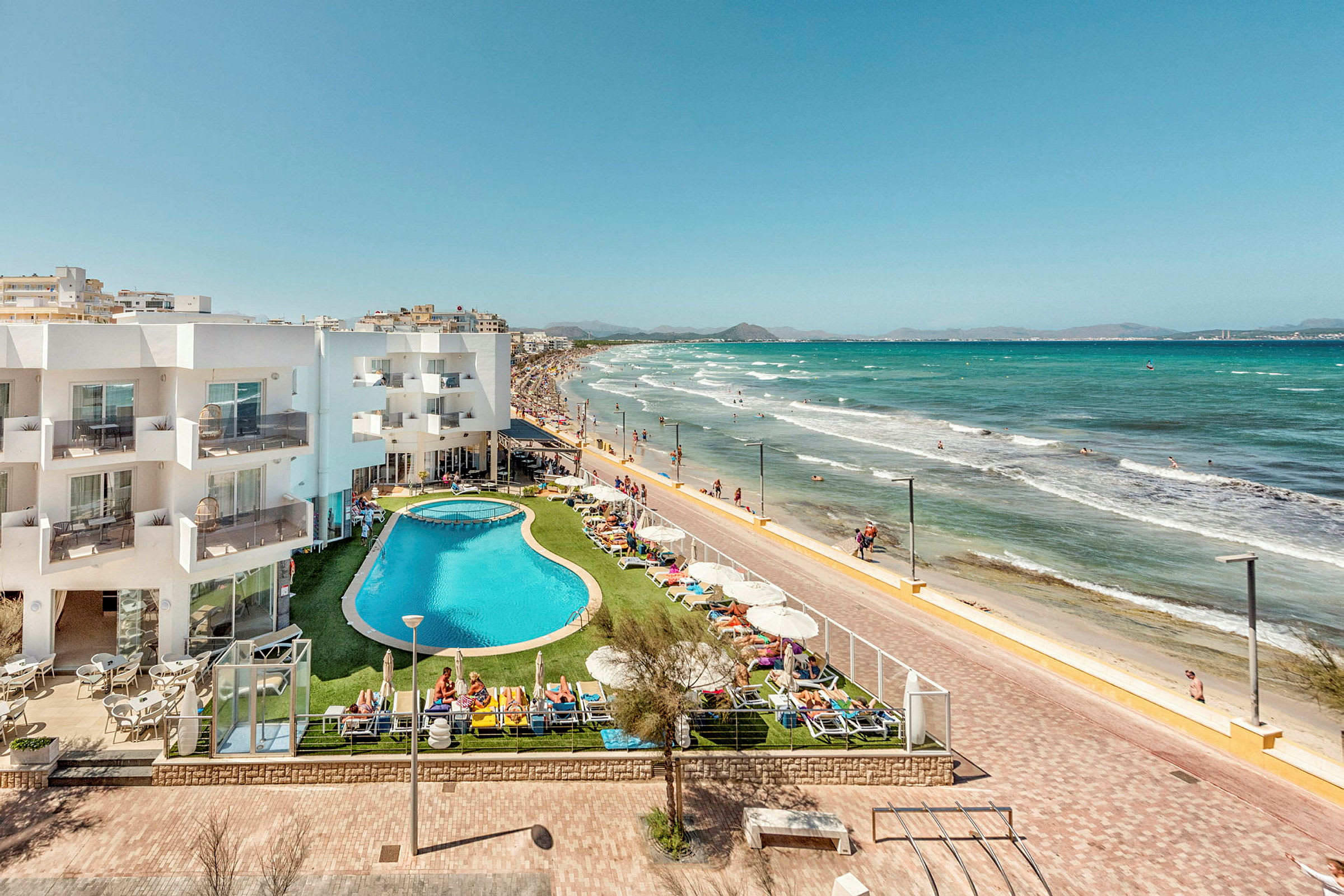 Hotellbyggnad och poolområde vid strandpromenad och sandstrand. (AI-genererad text)