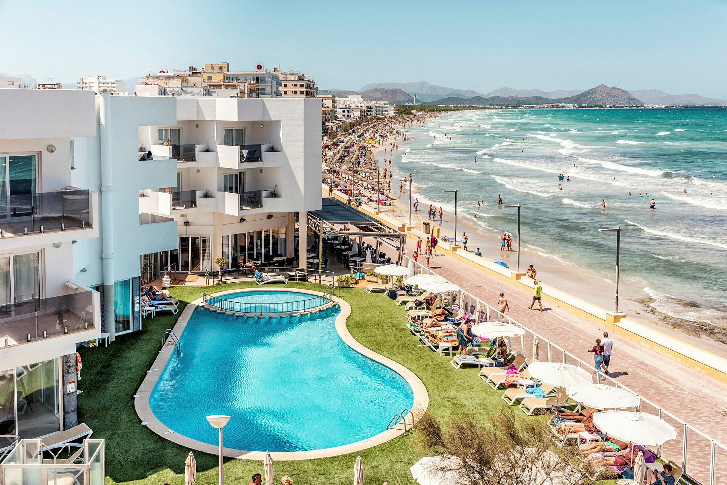 Bilden visar ett hotell med en pool nära stranden, omgiven av solstolar och parasoller. Strandpromenaden och havet syns i bakgrunden. (AI-genererad text)