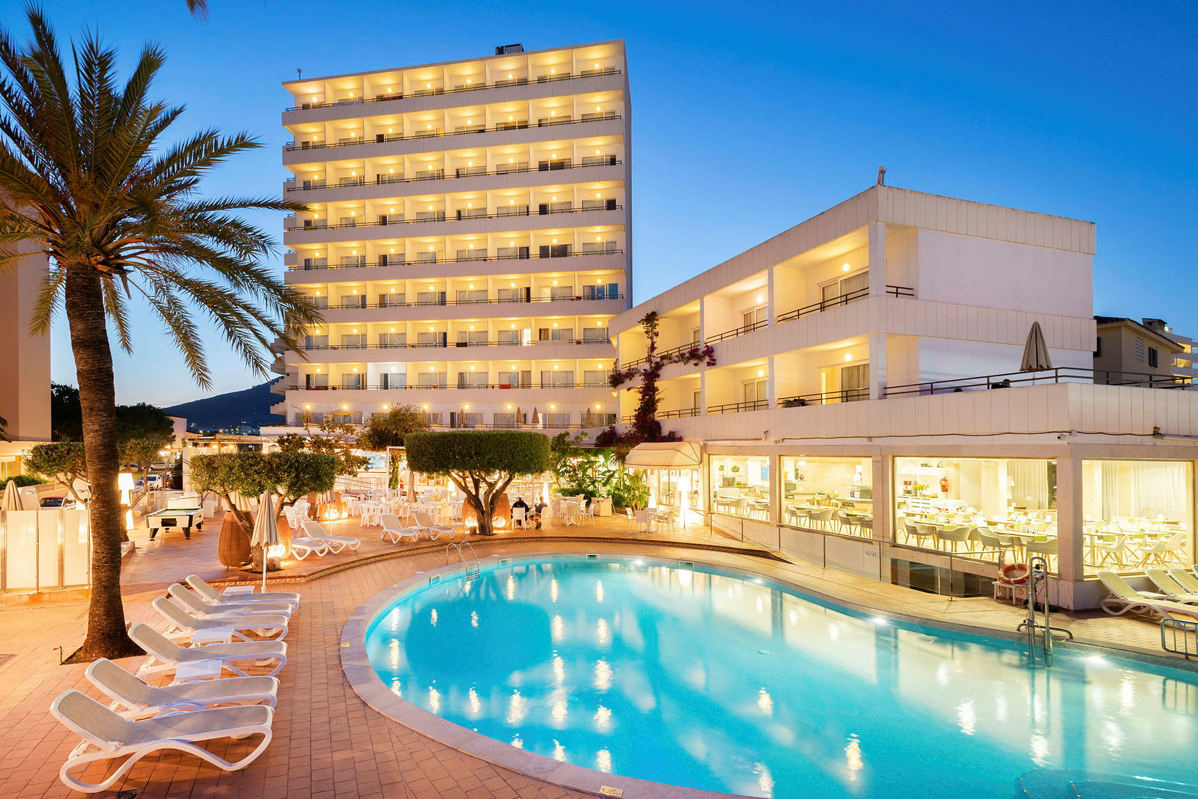 Hotellbyggnad med upplysta balkonger, en swimmingpool och solstolar vid skymning. (AI-genererad text)