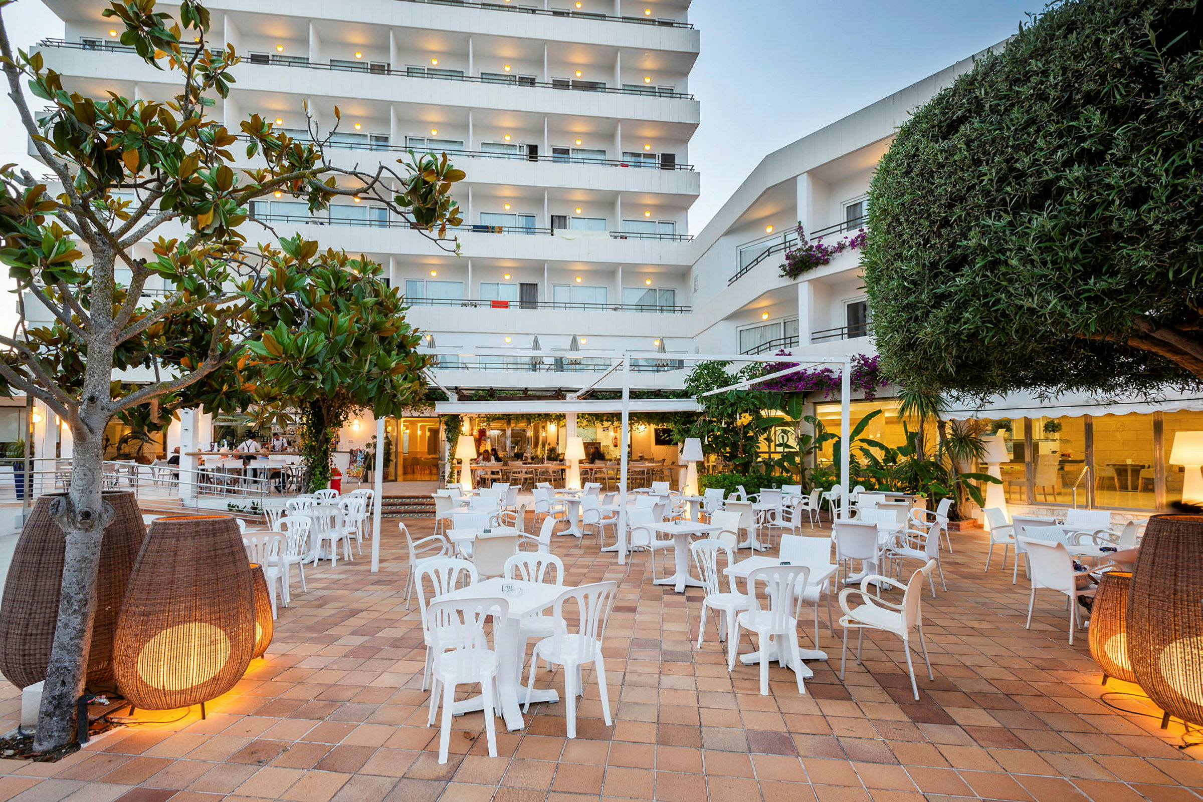 Utomhusområde med flera vita stolar och bord, omgivna av växter och modern belysning, med ett fler våningshotell i bakgrunden. (AI-genererad text)