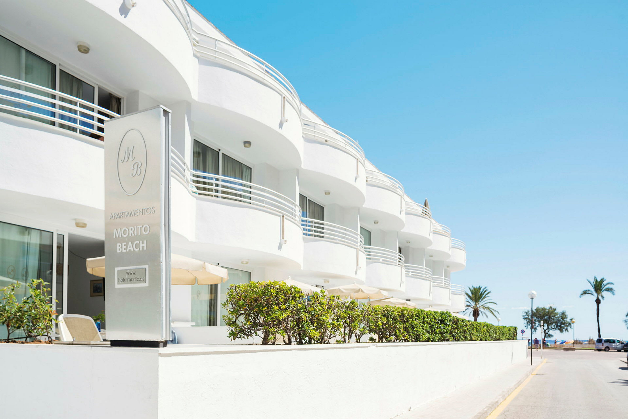 Den vita byggnaden tillhör Morito Beach Apartments, med palmer och hav i bakgrunden. (AI-genererad text)