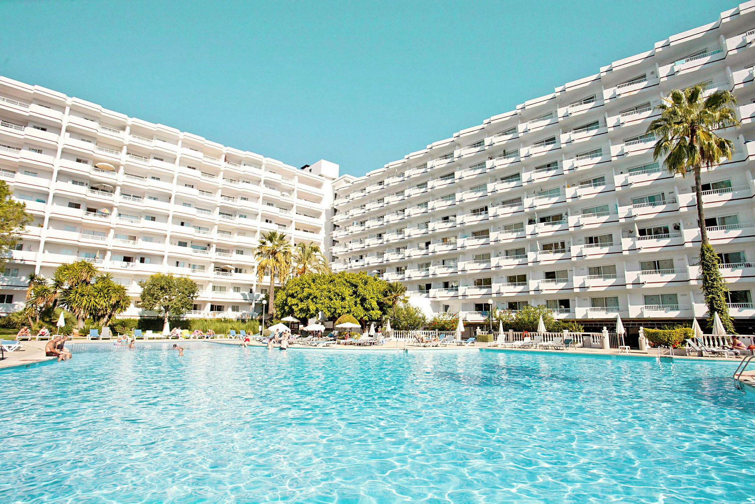 En stor pool omgiven av solstolar med ett hotell i bakgrunden. (AI-genererad text)