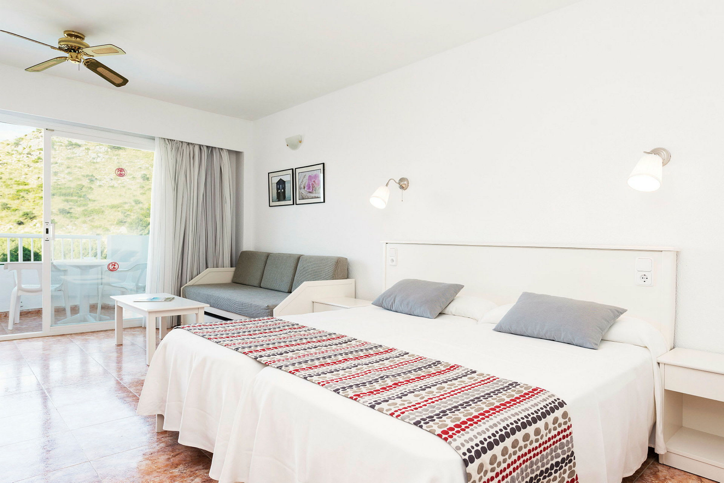 Ett ljust hotellrum med vita väggar och en dubbelsäng, en soffa, ett bord, och en balkongdörr som leder till en terrass med utsikt över grönska. (AI-genererad text)