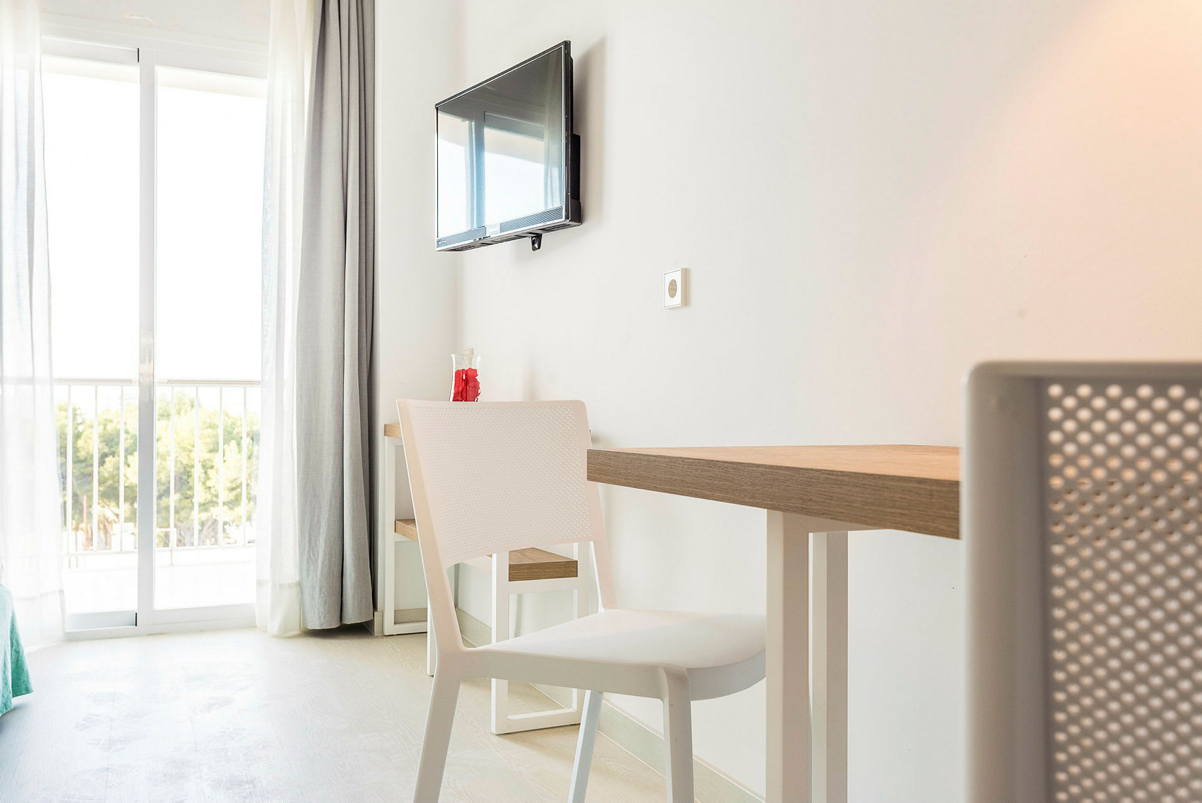 Ett hotellrum med vit inredning, ett bord och stolar samt en väggmonterad TV. I bakgrunden syns glasdörrar som leder till en balkong med utsikt över grönska. (AI-genererad text)