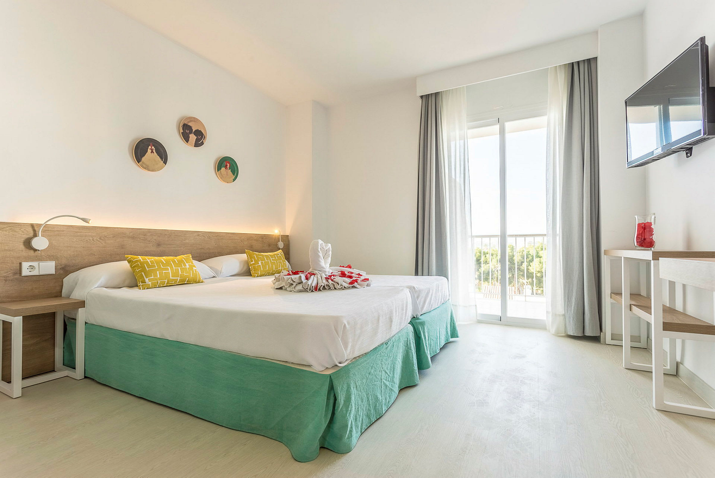 Ett hotellrum med två enkelsängar klädda i vitt sänglinne och gröna sängkjolar. Sängen är dekorerad med handdukskonst och röda blomblad. Det finns en vit vägg med dekorationer, ett skrivbord med en vas och en väggmonterad tv. Rummet har stora fönster med ljusa gardiner och utsikt mot träd. (AI-genererad text)