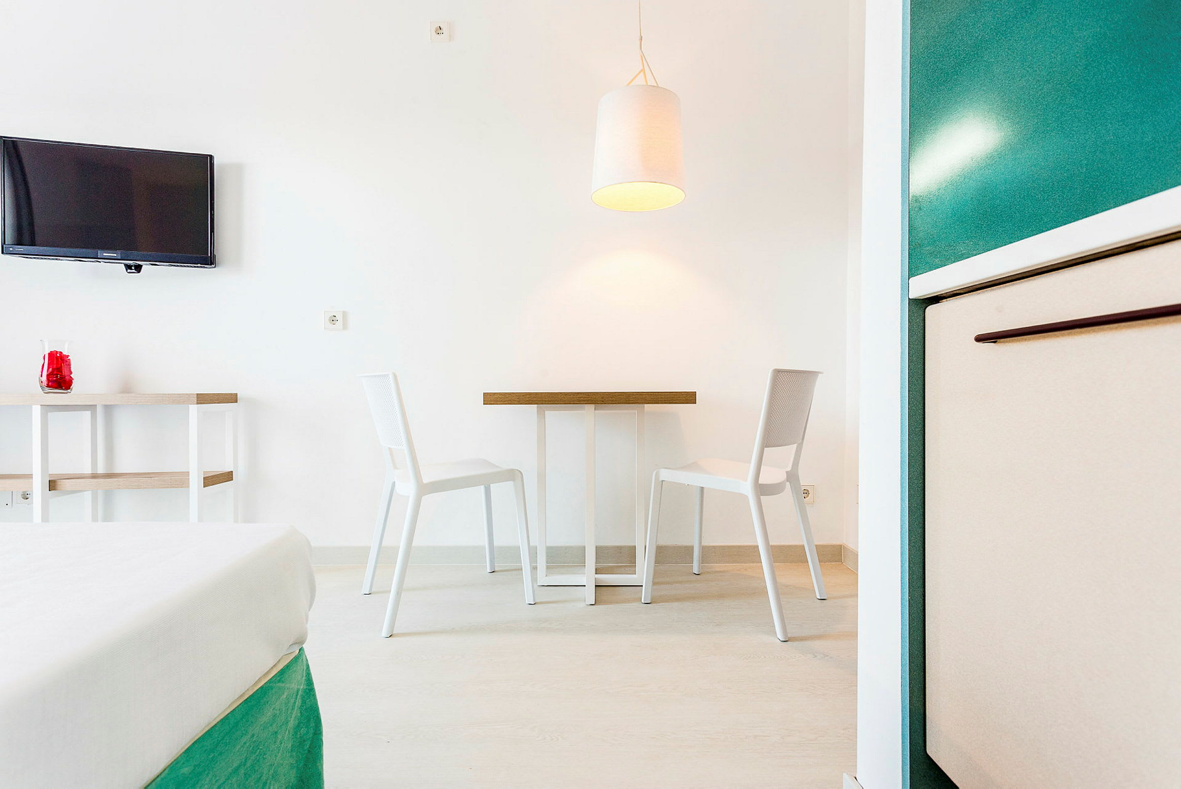 Ett rum med två vita stolar framför ett litet bord, en taklampa hänger ovanför bordet, och en TV är monterad på väggen till vänster. (AI-genererad text)