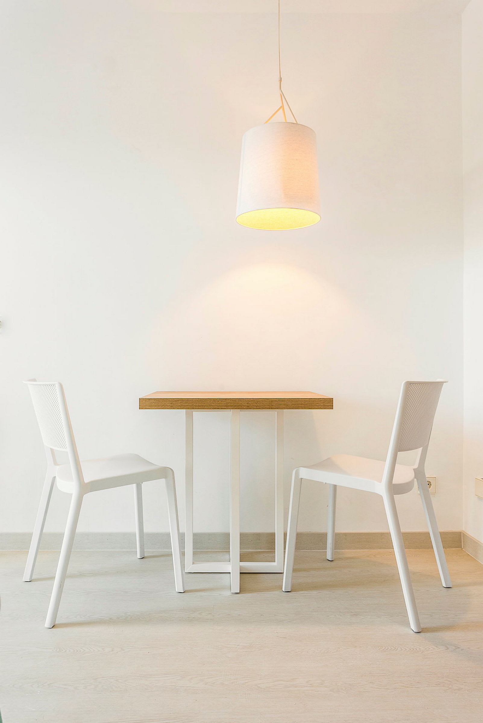 Ett bord med två vita stolar, och en hängande lampa ovanför i ett minimalistiskt rum. (AI-genererad text)