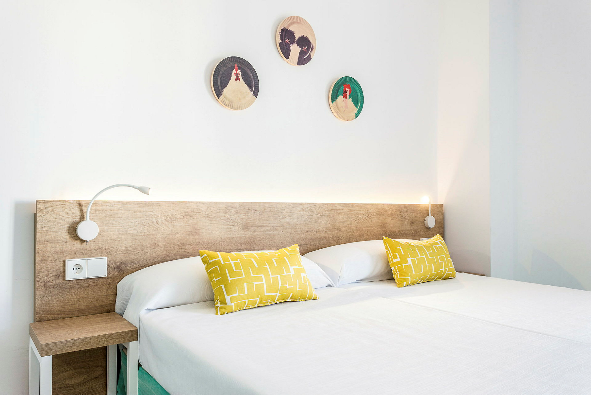 Ett modernt hotellrum med en dubbelsäng. Sängen har en träbas, vita lakan och två gula kuddar. Väggdekorationer med mönster av fåglar är uppsatta ovanför huvudänden. (AI-genererad text)