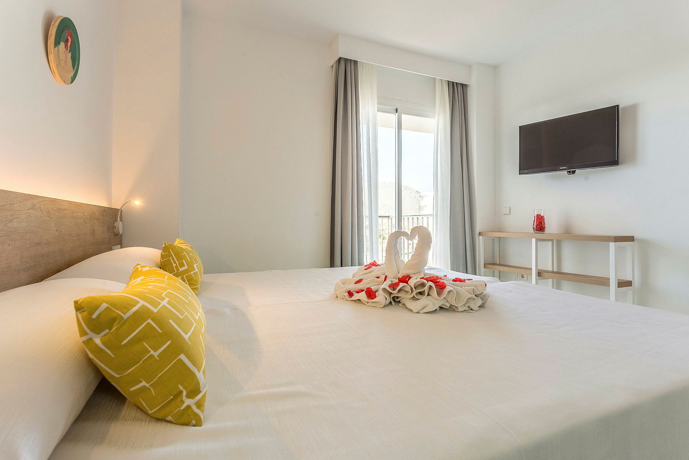 Ett hotellrum med en stor säng, dekorerad med handduksvikningar och blommor. Rummet har en TV på väggen och ett fönster med gardiner. (AI-genererad text)