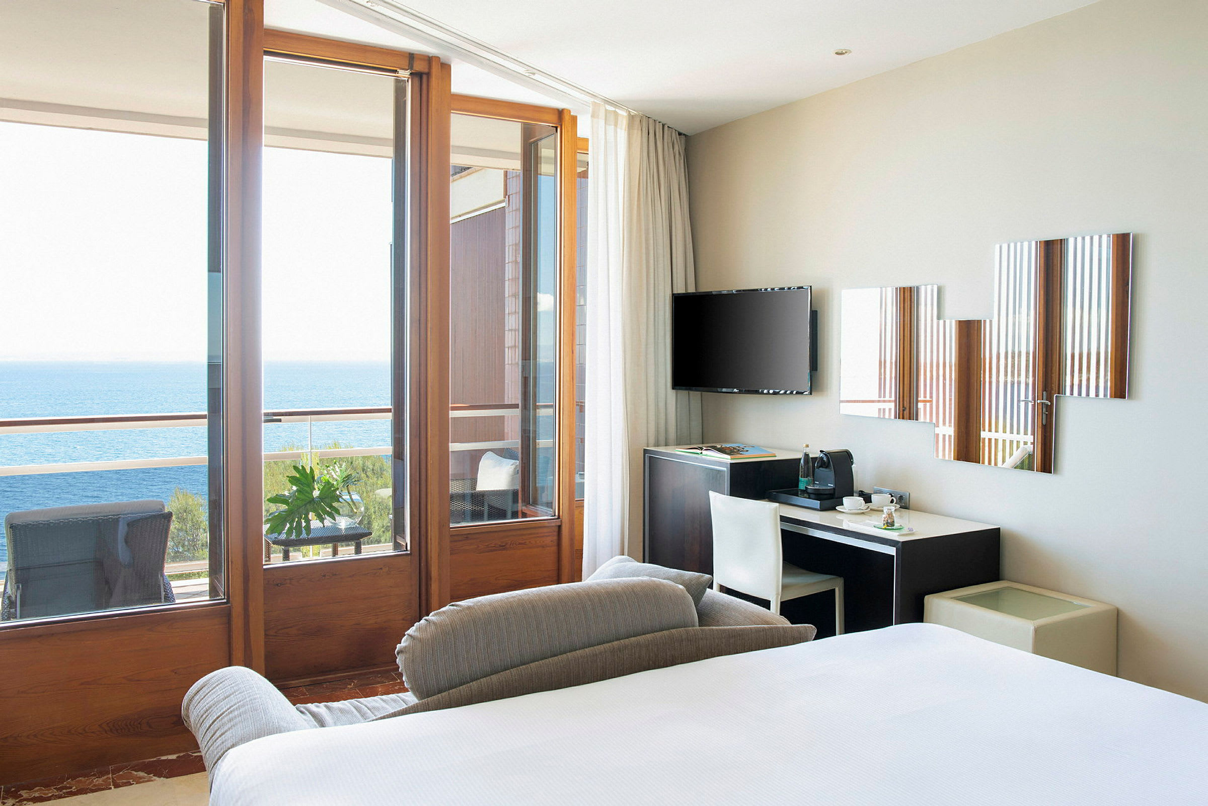 En modern hotellrumsvy med en säng, skrivbord, TV, och stora glasdörrar som leder ut till en balkong med utsikt över havet. (AI-genererad text)