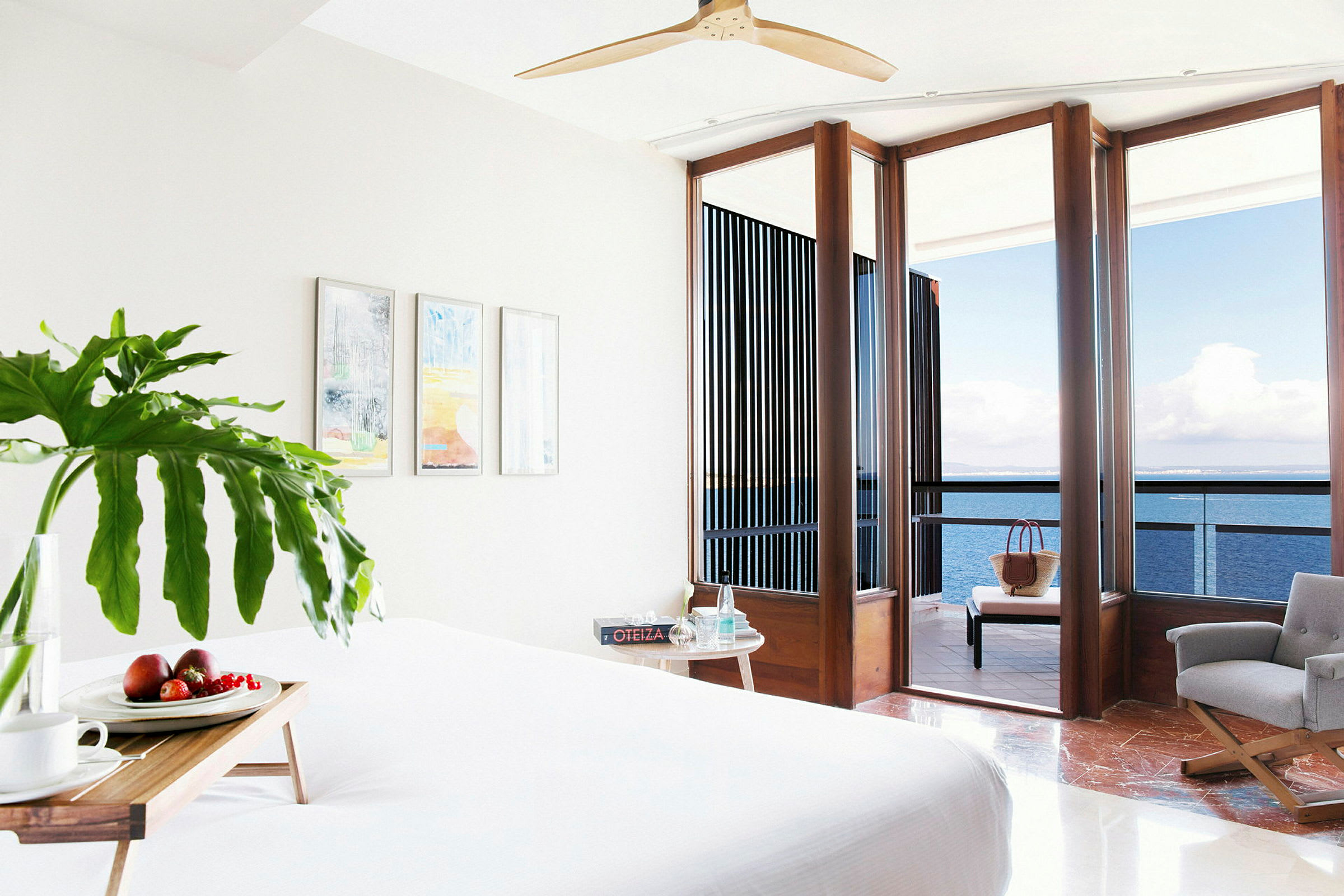 Ett hotellrum med vit säng, färska frukter på ett träbricka, stora fönster och balkong med utsikt över havet. (AI-genererad text)