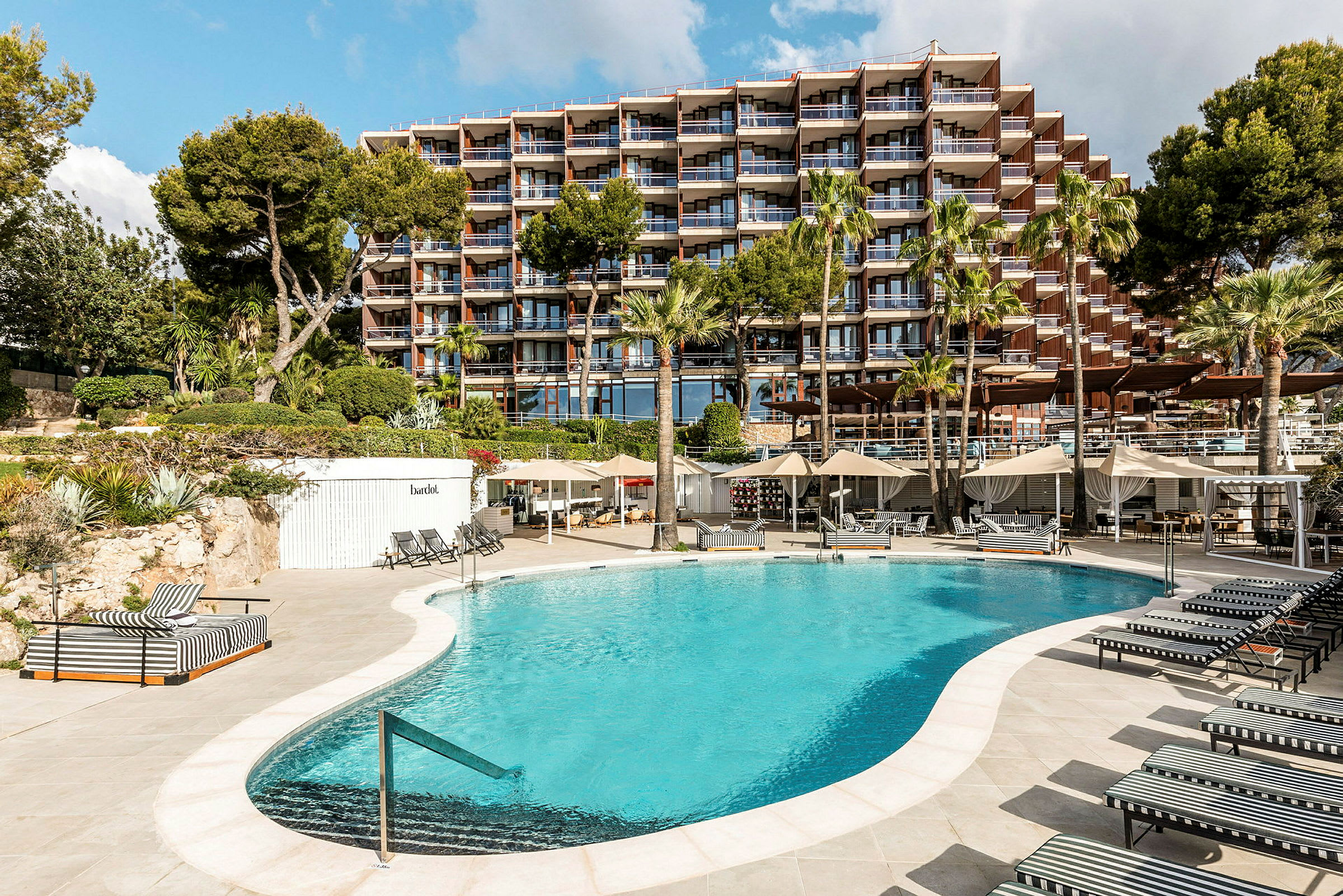 En utomhuspool omgiven av solstolar och parasoller framför en större hotelbyggnad. (AI-genererad text)