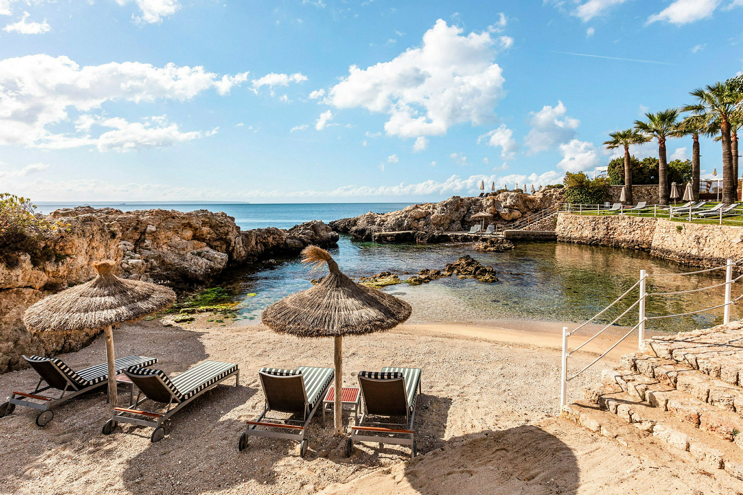Strand med halmtakssparasoller, solstolar och rofylld utsikt över havet och en liten klippig vik. (AI-genererad text)