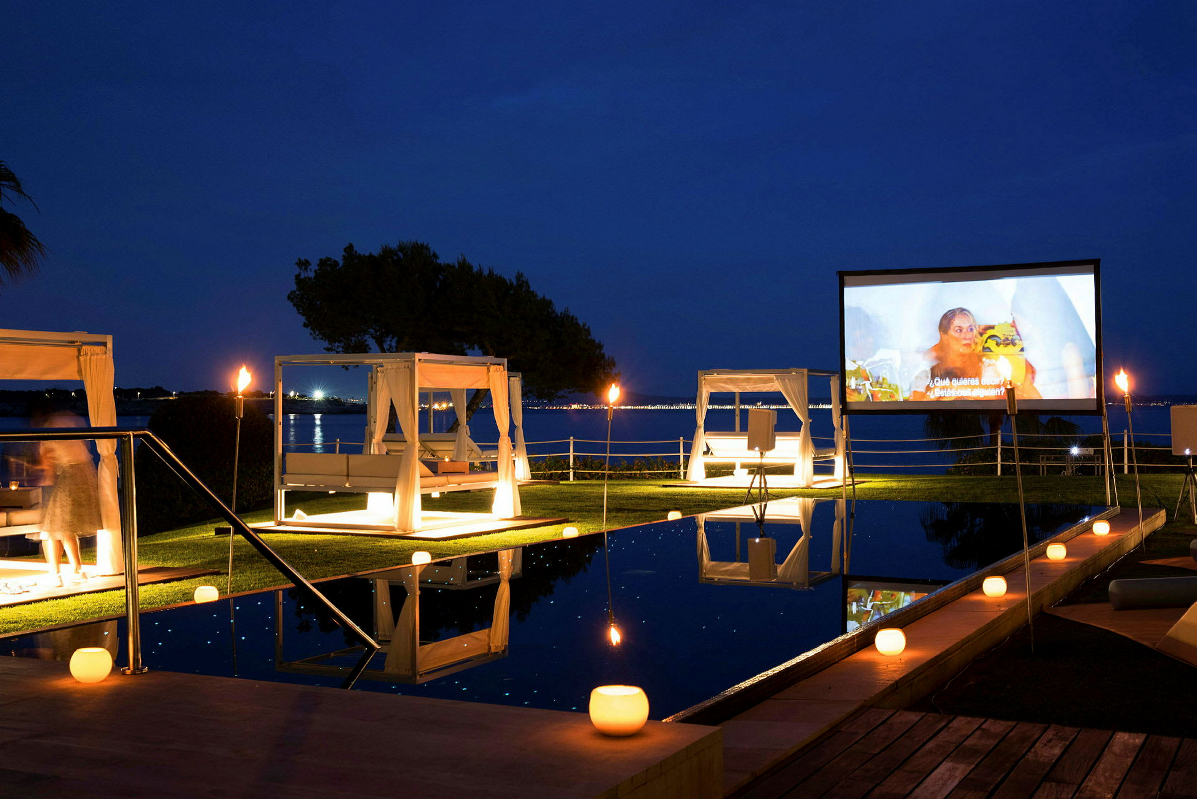 Uteplats med pool, vita himmelssängar och en stor filmduk nära vatten på kvällen. (AI-genererad text)