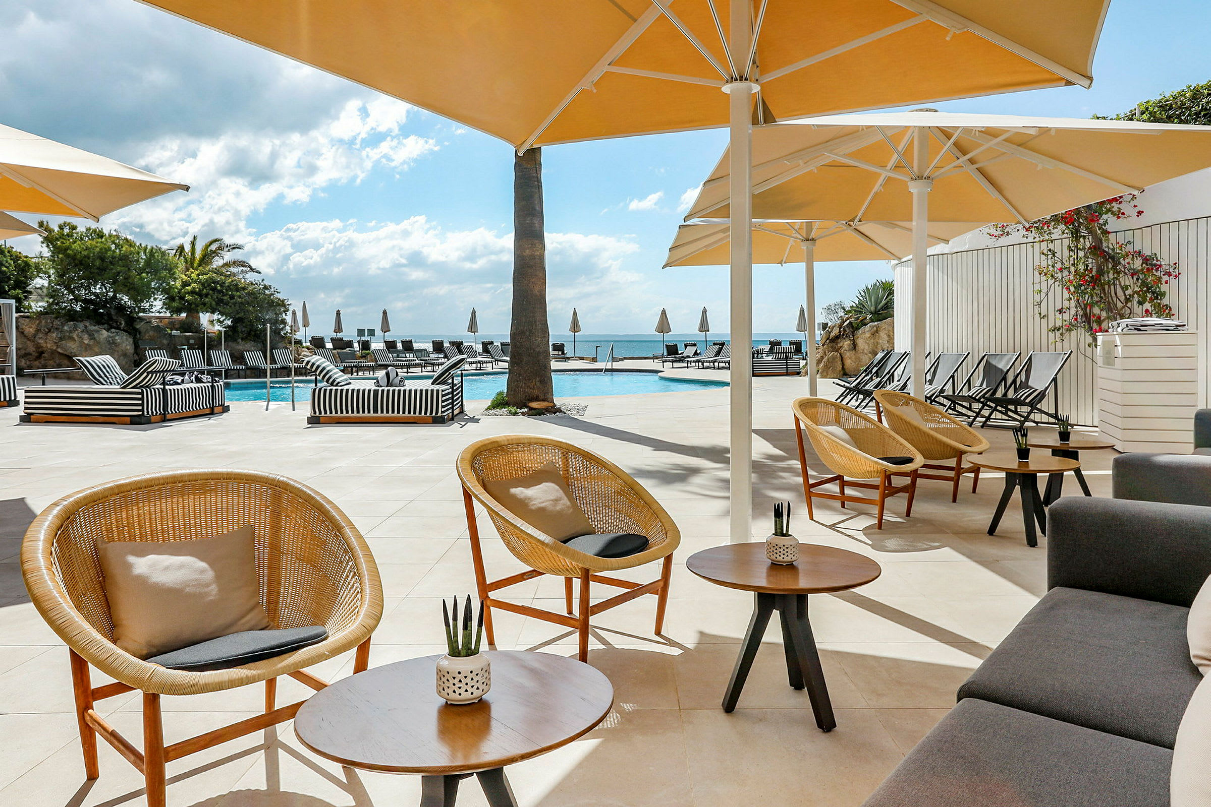 Bild av en poolterrass med parasoller, stolar och soffor. Ett klart blått himmel och träd i bakgrunden. (AI-genererad text)