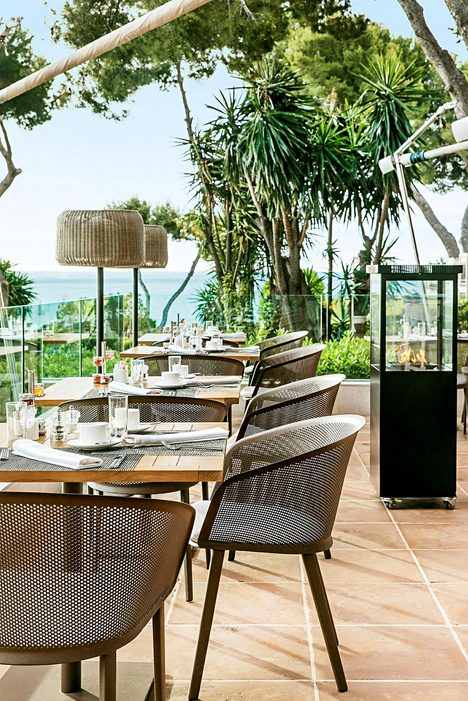 Utomhusrestaurang med moderna stolar och dukade bord, omgiven av grönska och utsikt mot havet. (AI-genererad text)