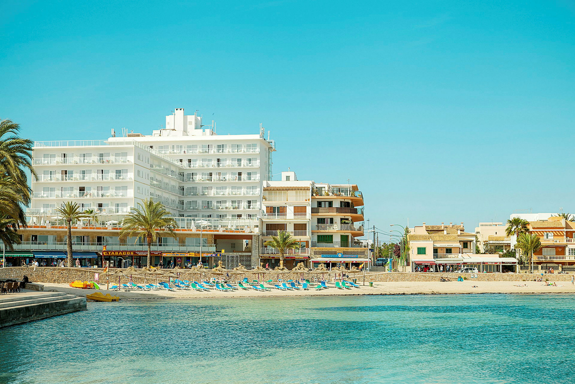 En strand med solsängar och parasoller framför ett område med hotell och byggnader. (AI-genererad text)