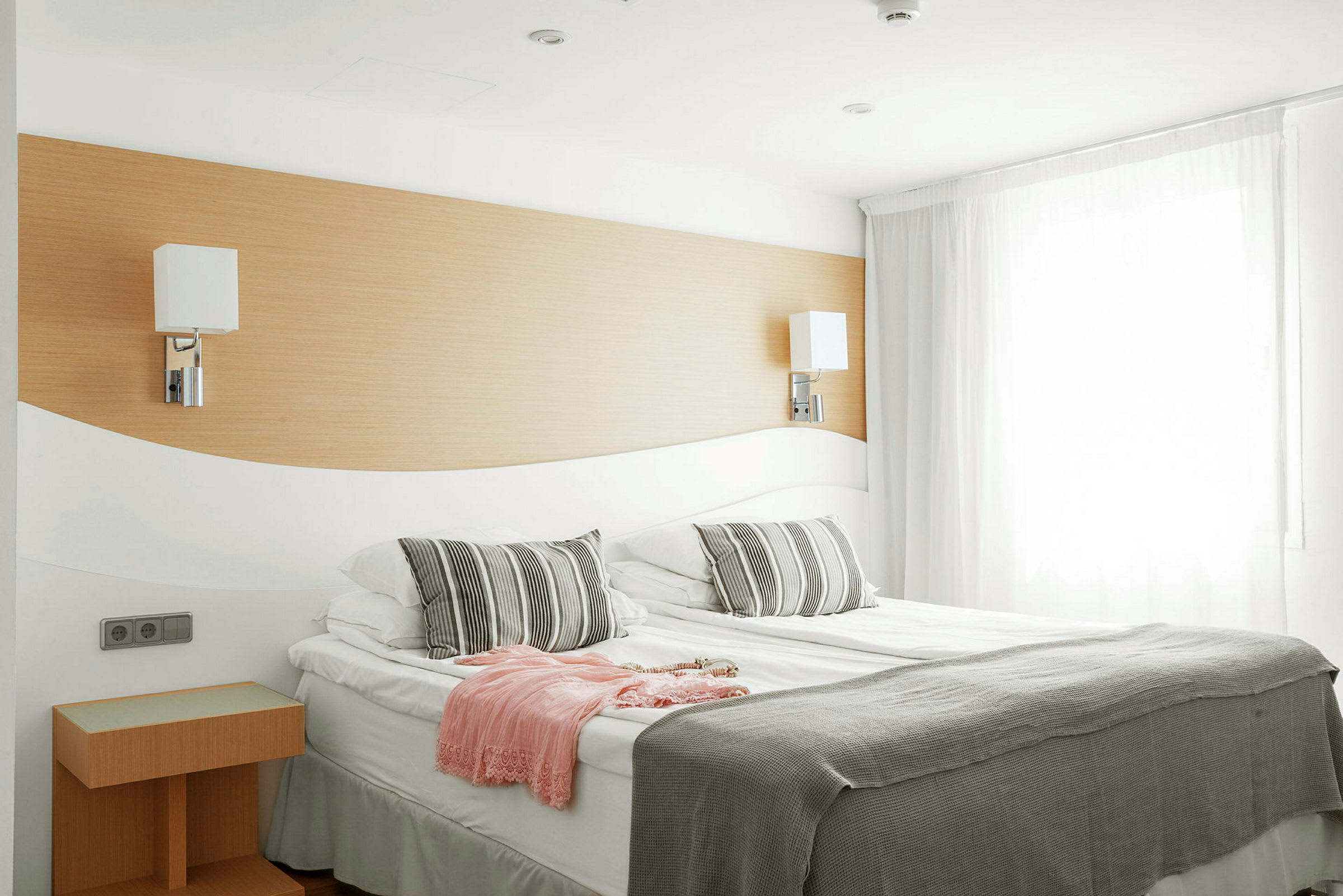 Ett hotellrum med en dubbelsäng, två lampor på väggen, och ett täcke samt kuddar i grått och vitt. (AI-genererad text)